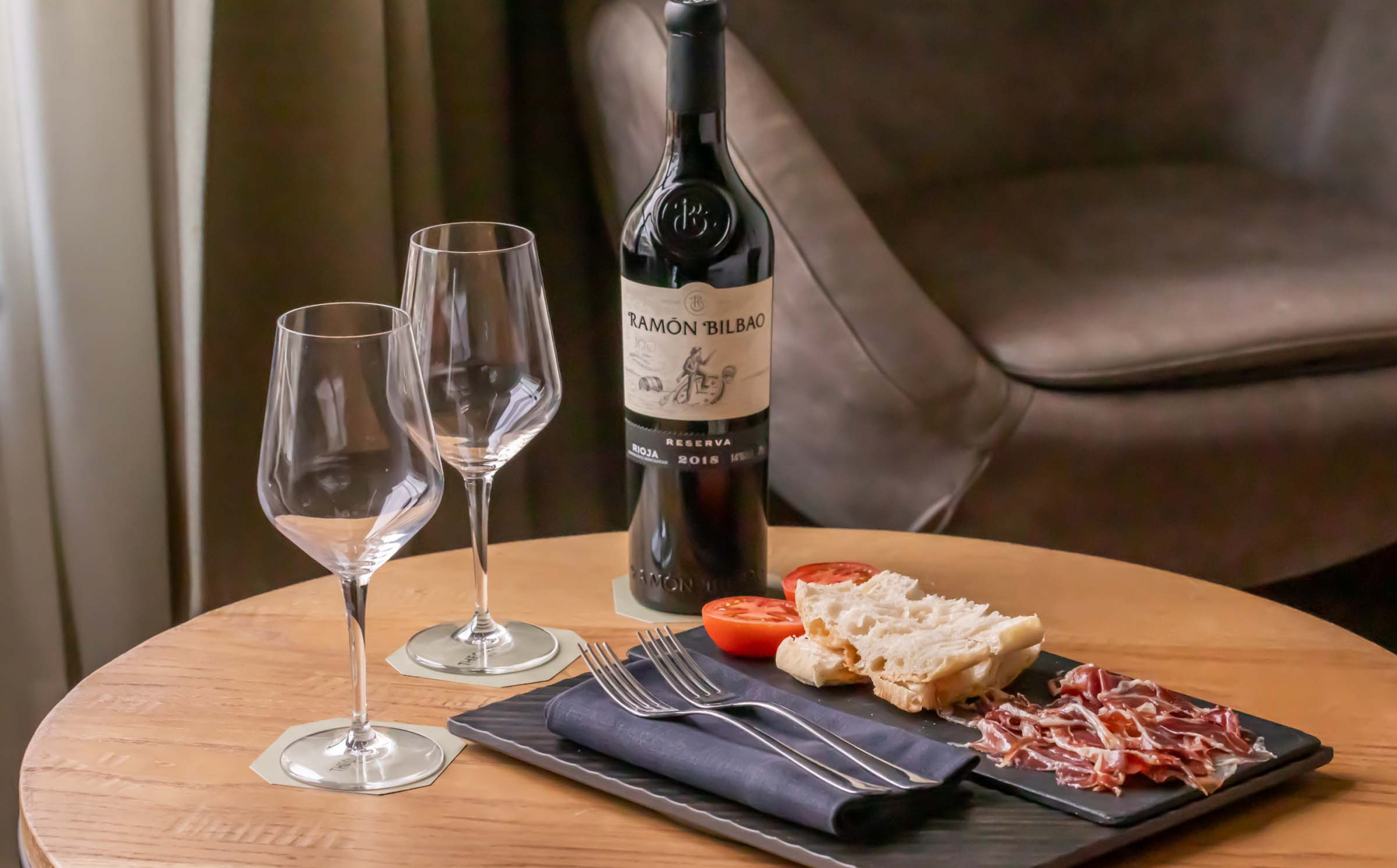 Sobre una mesa de madera se ve una botella de vino, dos copas y un plato con aperitivos y embutido.