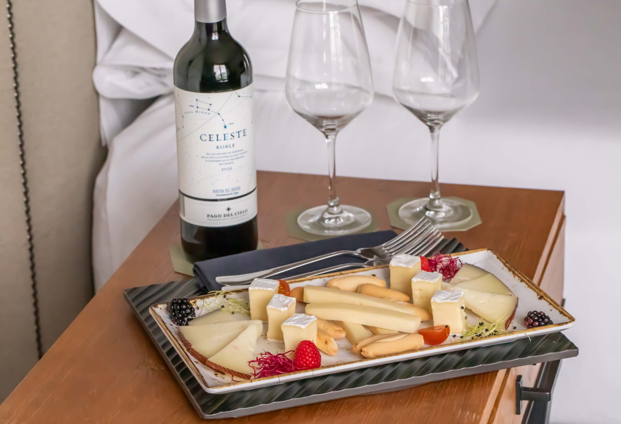 Una bandeja con queso y verduras junto a una botella de vino y copas de cristal sobre una mesa de madera.
