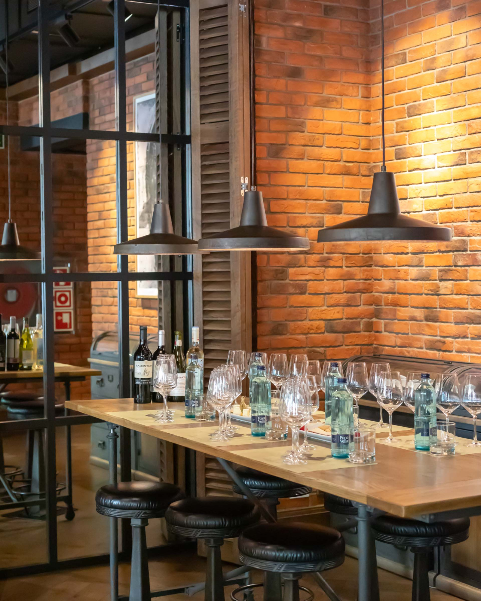 Presenta una barra con botellas, vasos y taburetes, junto a una pared de ladrillo y lámparas colgantes.
