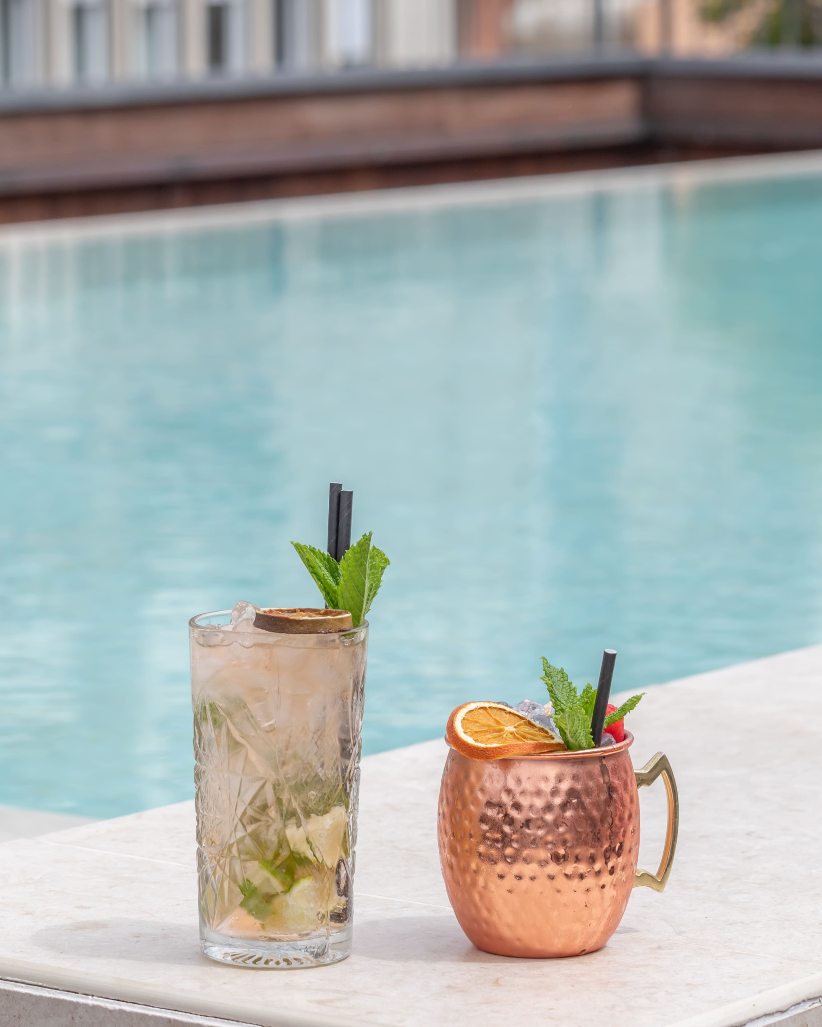 Se ve una mesa junto a una piscina exterior con dos bebidas servidas en vasos y una copa de color cobre.