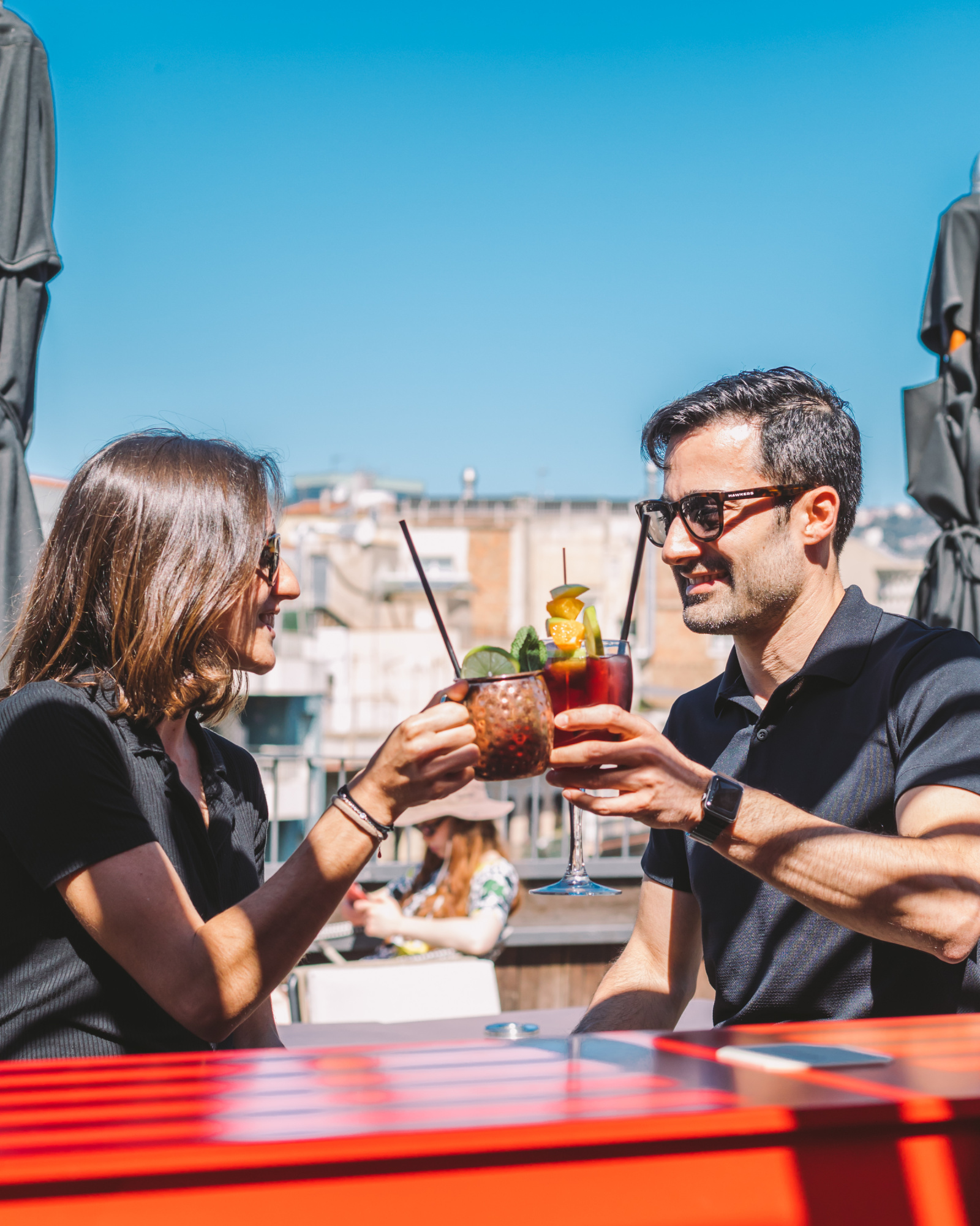 Dos personas brindan con bebidas en una terraza con sillas y sombrillas bajo el cielo abierto.