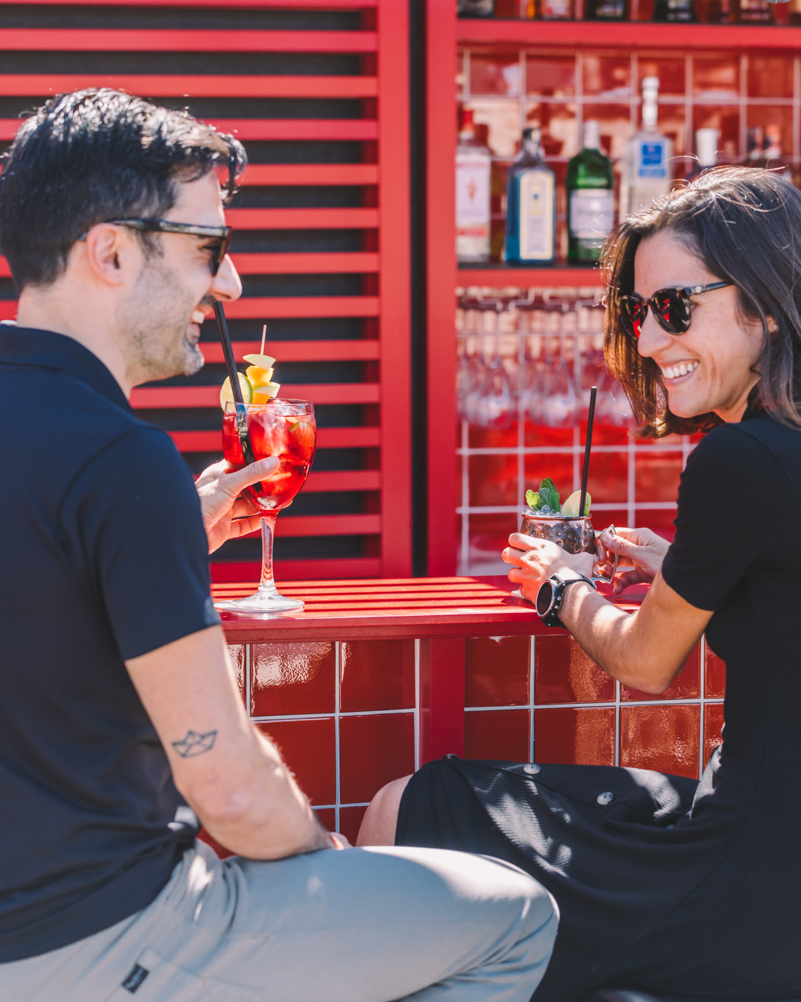 Dos personas disfrutan de cócteles en una barra con botellas y vasos detrás.