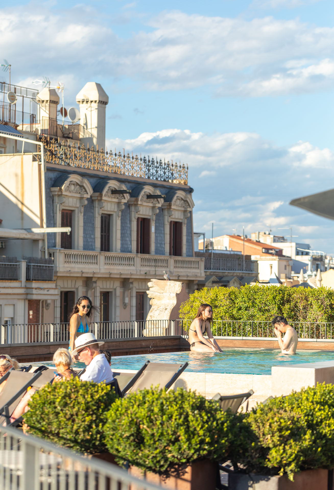 Varias personas disfrutan de la piscina exterior junto a plantas y barandillas en una terraza al aire libre.