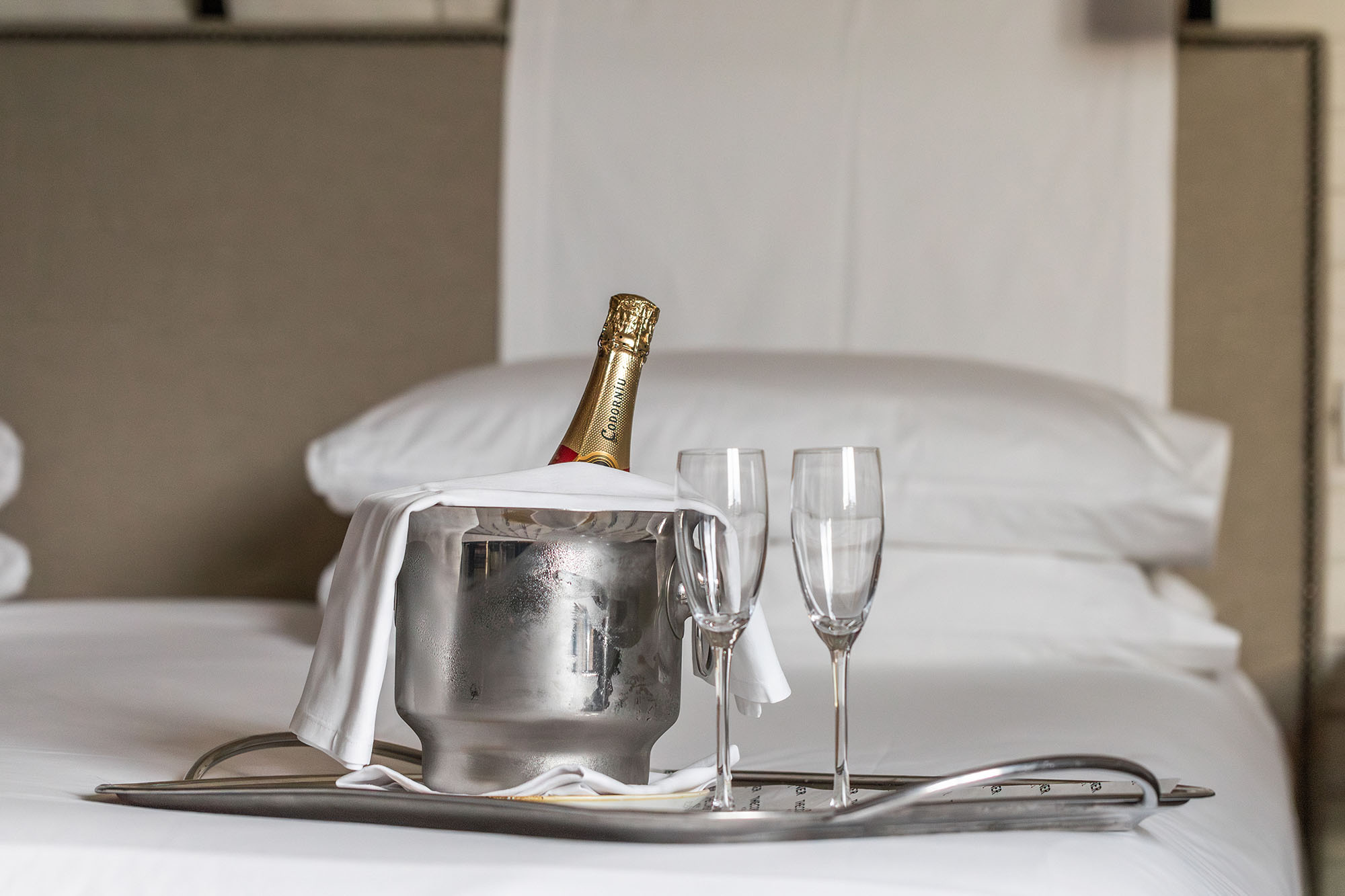 Se ve una cubitera con botella de cava y copas sobre una bandeja en una cama con sábanas blancas.