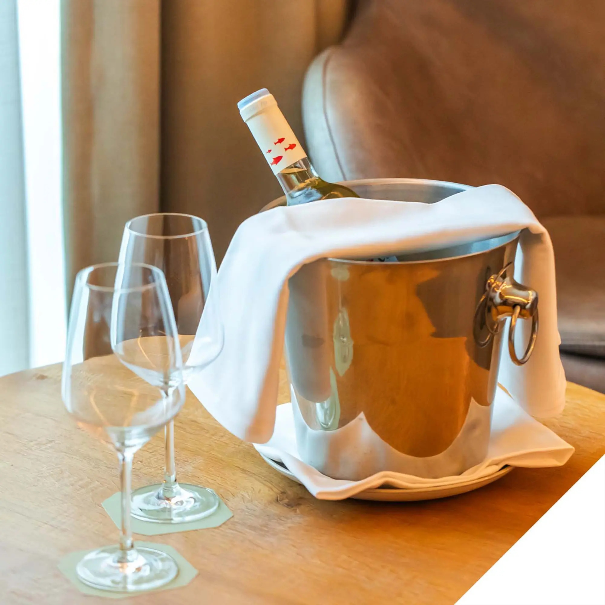 Se ve una cubitera plateada con una botella de vino sobre una mesa junto a dos copas.