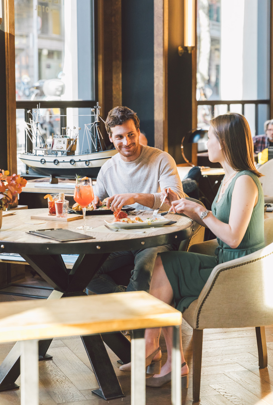 Due persone conversano e mangiano a un tavolo accanto a grandi finestre in un’area ristorante.