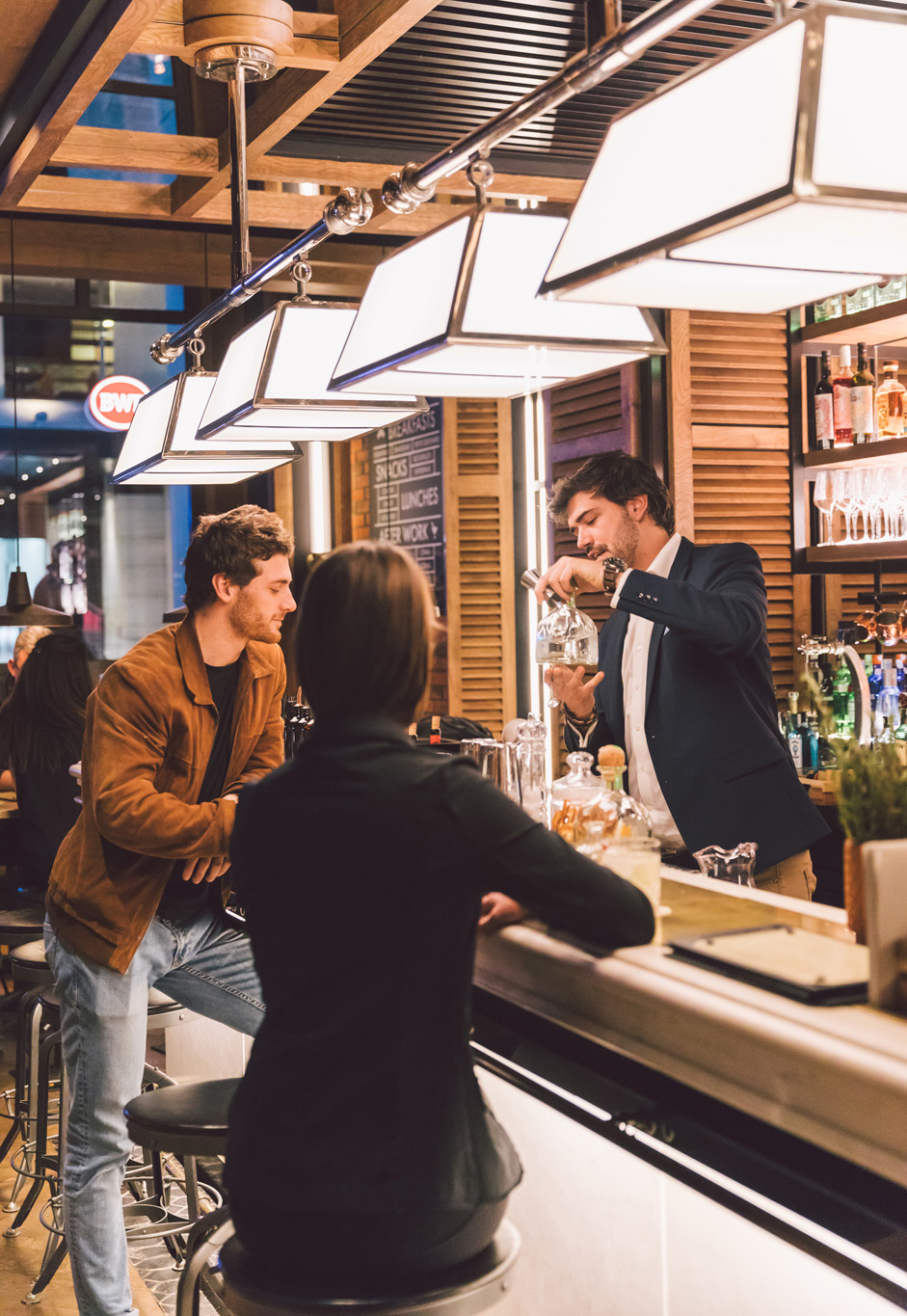 Diverse persone chiacchierano e bevono a un bar illuminato, con bicchieri e bottiglie ben visibili.