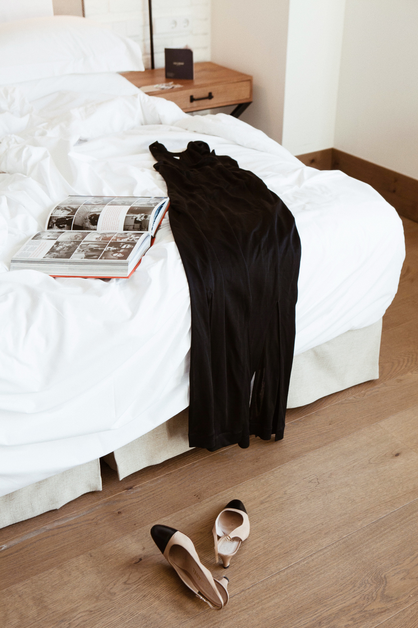 Se ve una cama con ropa de cama blanca y una mesita de noche de madera con lámpara y objetos sobre ella.