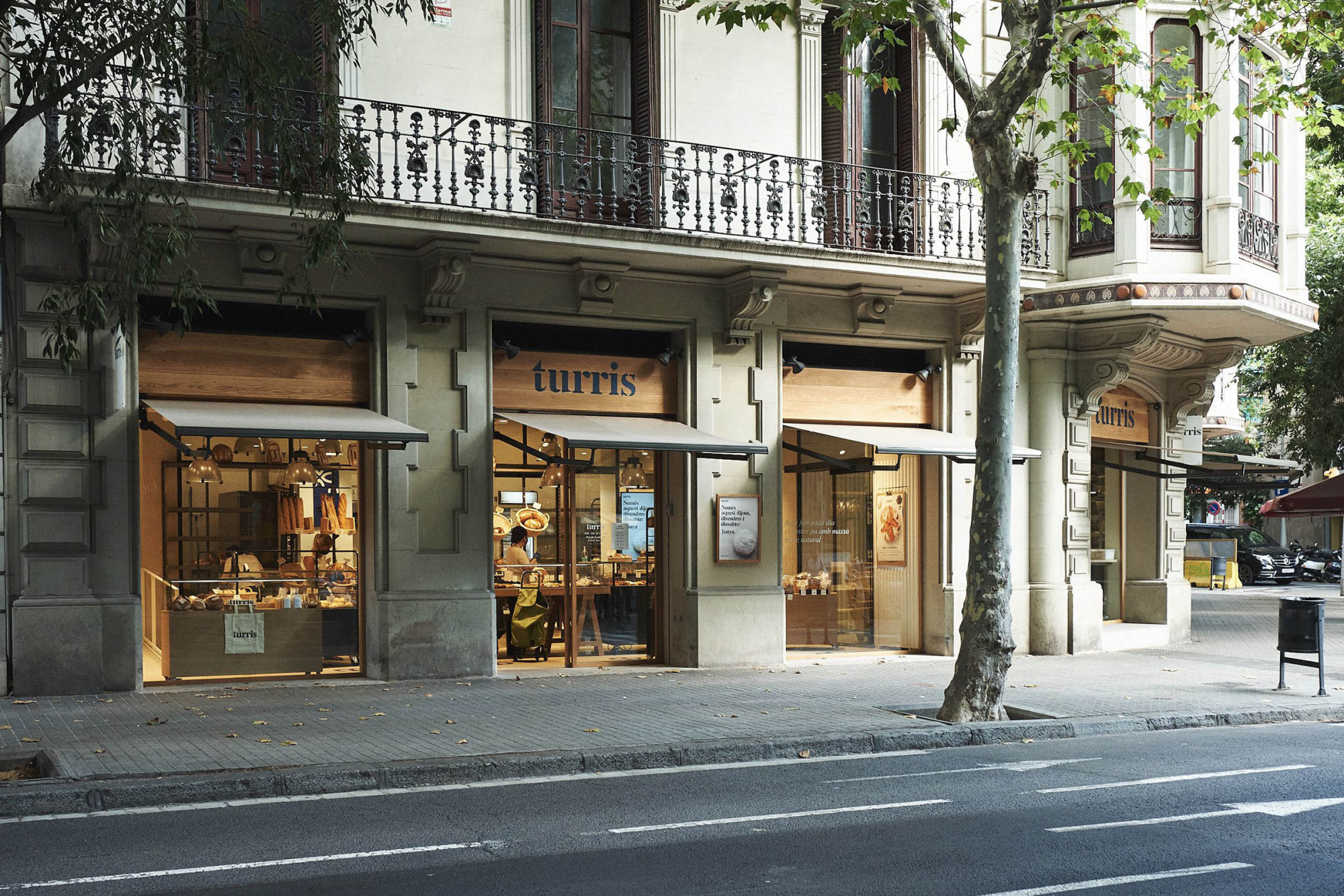 Se ve una fachada de edificio con balcones, árboles en la acera y dos escaparates a pie de calle.