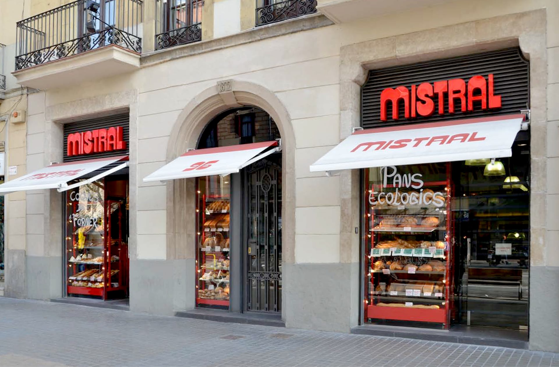 La imagen muestra una fachada de edificio con tres escaparates y toldos rojos de “MISTRAL” en una calle peatonal.