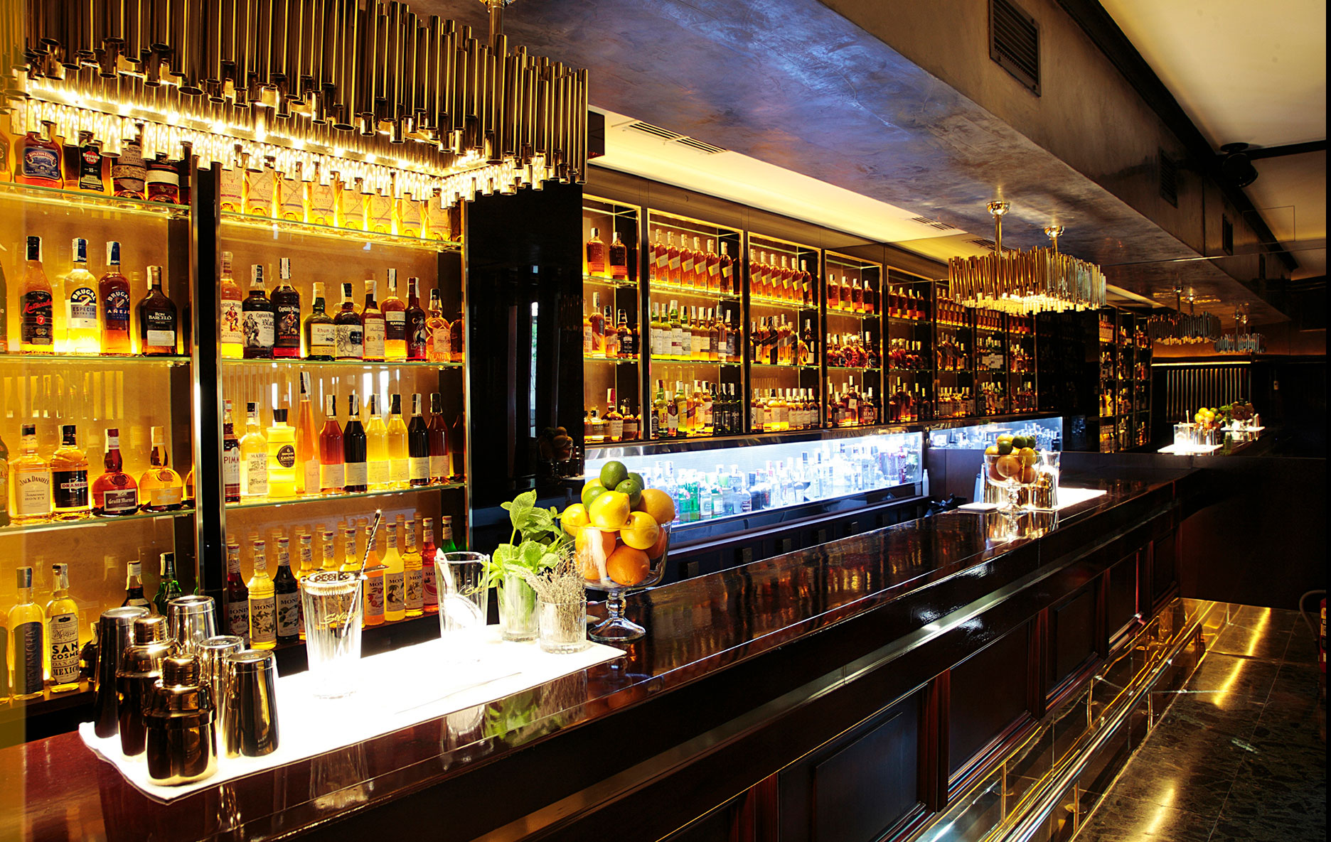 El bar muestra una barra larga con botelleros de vidrio retroiluminados y vasos sobre la encimera.