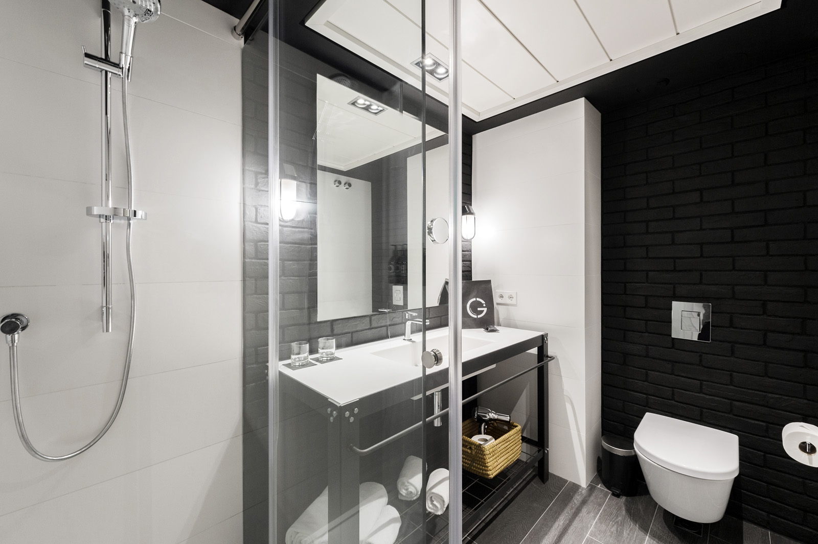 Baño moderno con ducha de cristal, lavabo con encimera blanca e inodoro junto a una pared de azulejos oscuros.