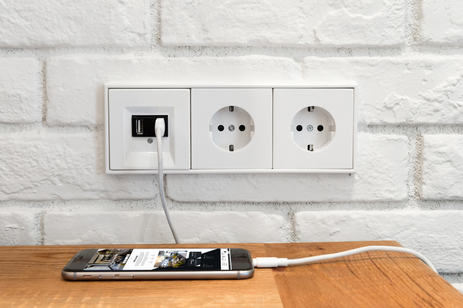 Se ve una toma de corriente múltiple empotrada en una pared de ladrillo blanco con un teléfono sobre una mesa de madera.