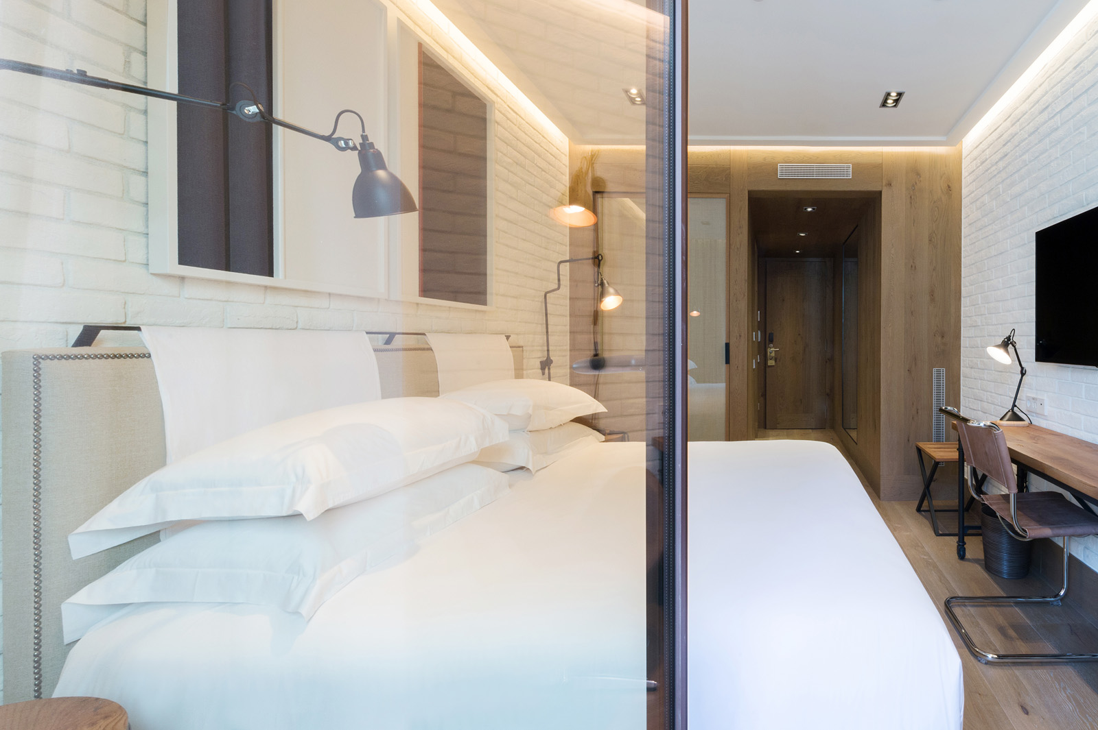 Se ve una cama doble con ropa de cama blanca, una lámpara junto a la pared y un escritorio en el interior.