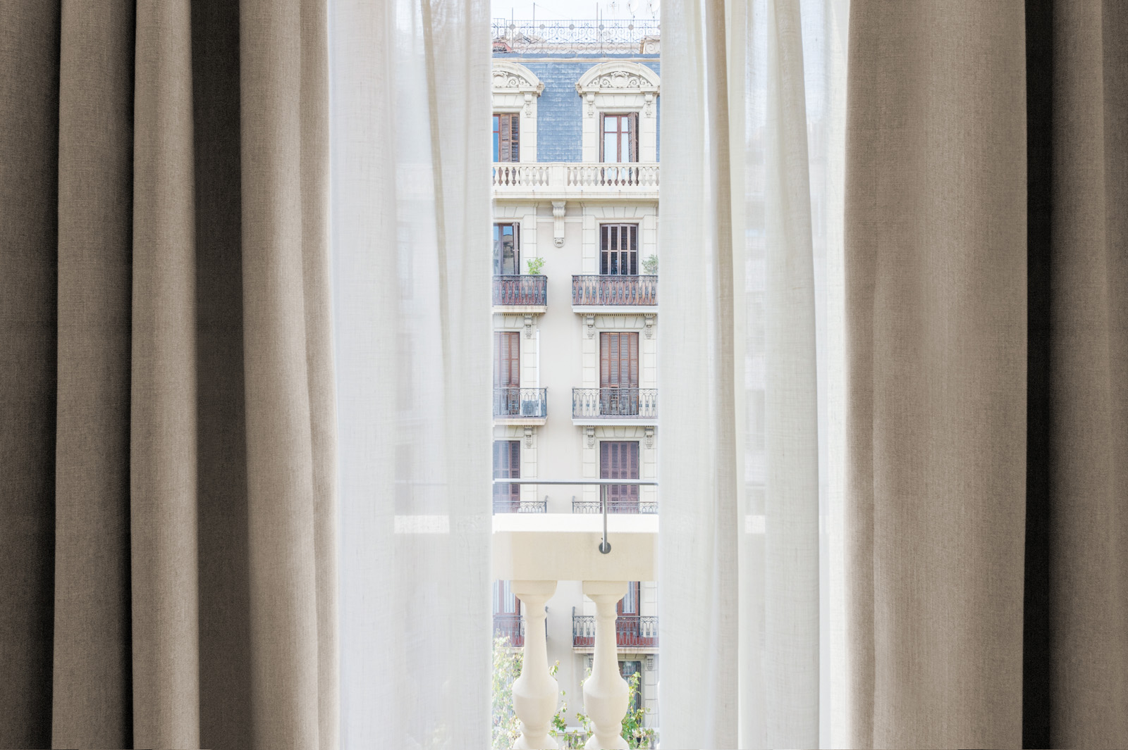 Se ve una ventana con cortinas claras abiertas dejando entrar luz natural y mostrando balcones del edificio de enfrente.