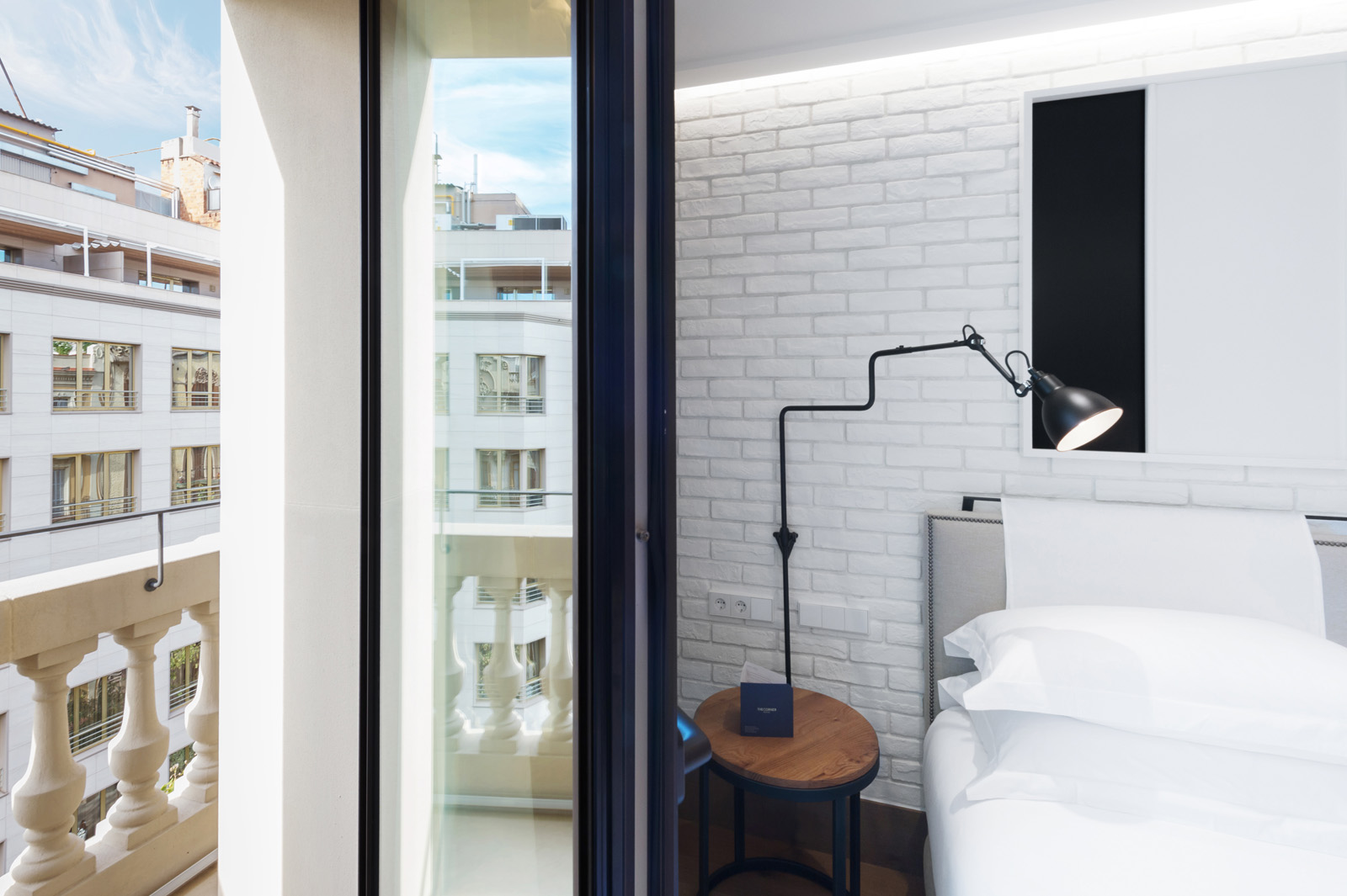 Se ve una zona de descanso con cama blanca, lámpara de pie y puerta corredera hacia un balcón con vista al exterior del edificio.