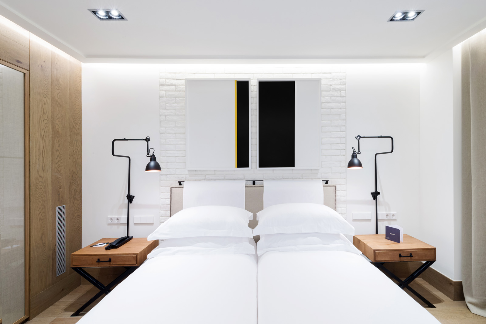 La imagen muestra una cama con ropa de cama blanca y mesitas de noche junto a dos lámparas de pared en una habitación luminosa.