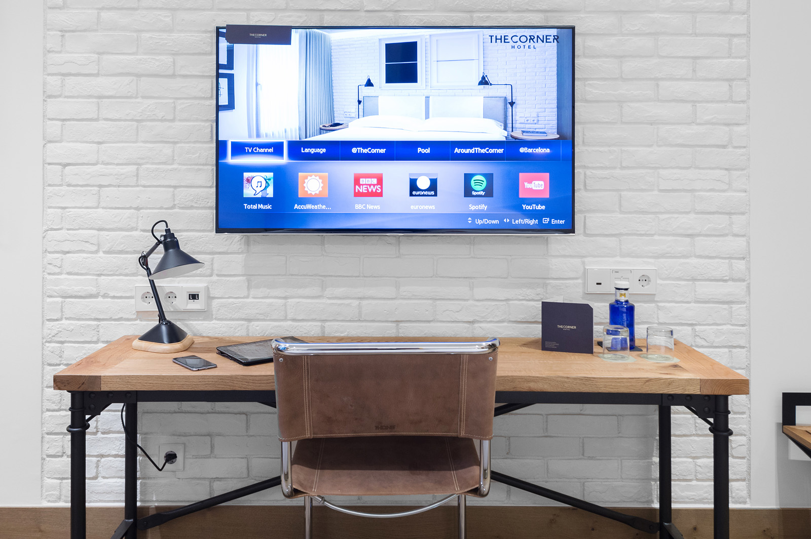 Se ve un escritorio con silla y un monitor sobre una pared de ladrillo con lámpara y artículos sobre la mesa.