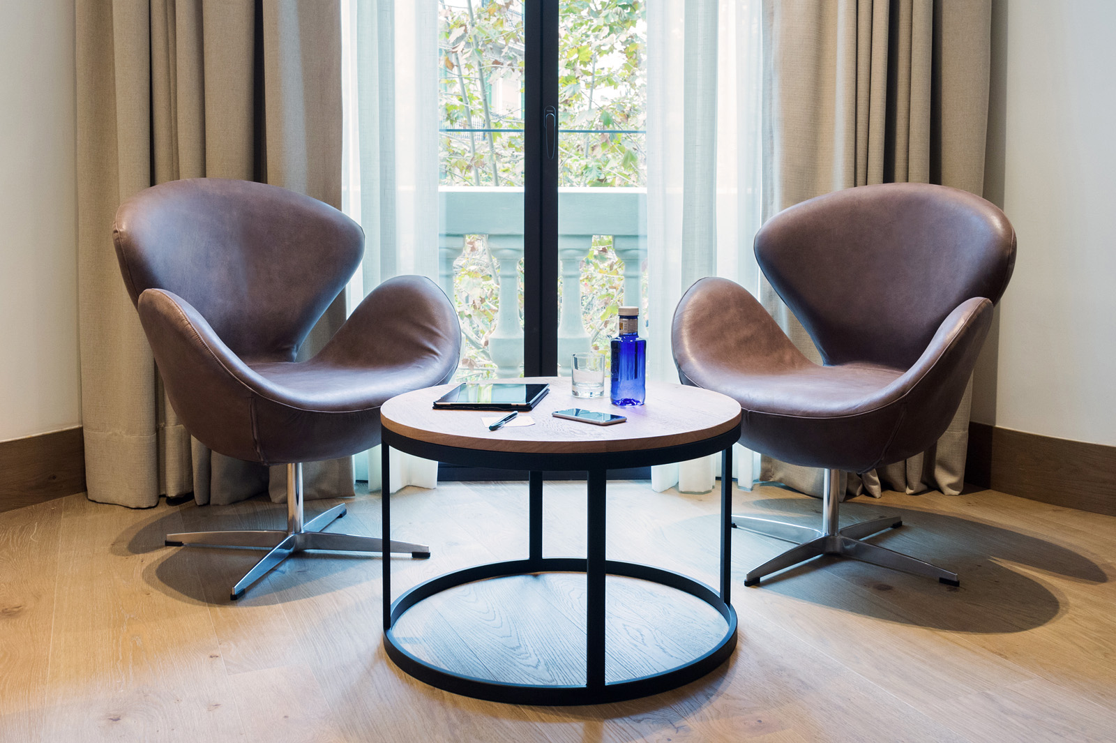 Presenta dos sillones tapizados y una mesa redonda con una zona de asientos frente a una ventana con cortinas.