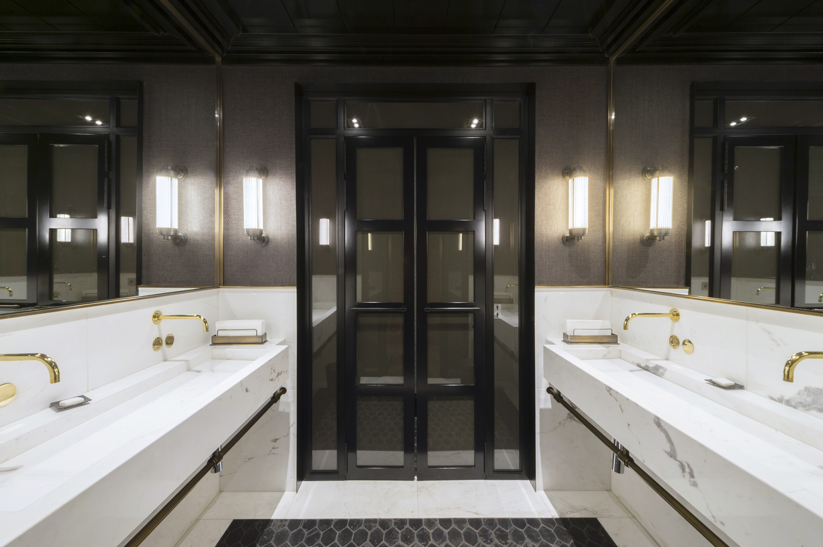 Baños modernos con dos lavabos, encimera blanca y espejo iluminado sobre puertas de vidrio.