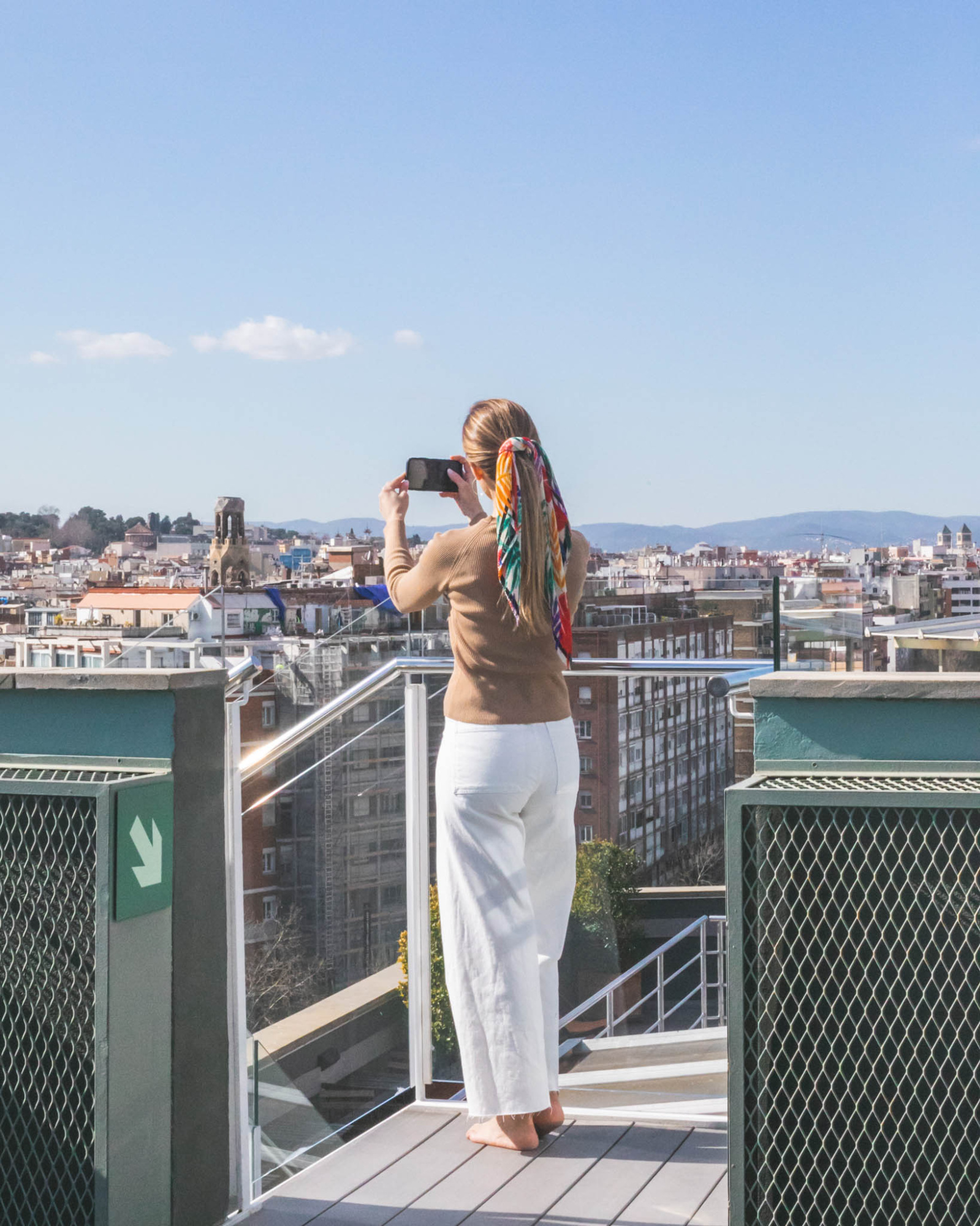 Una mujer camina sobre una terraza junto a una barandilla metálica, con una piscina rectangular y la ciudad al fondo.