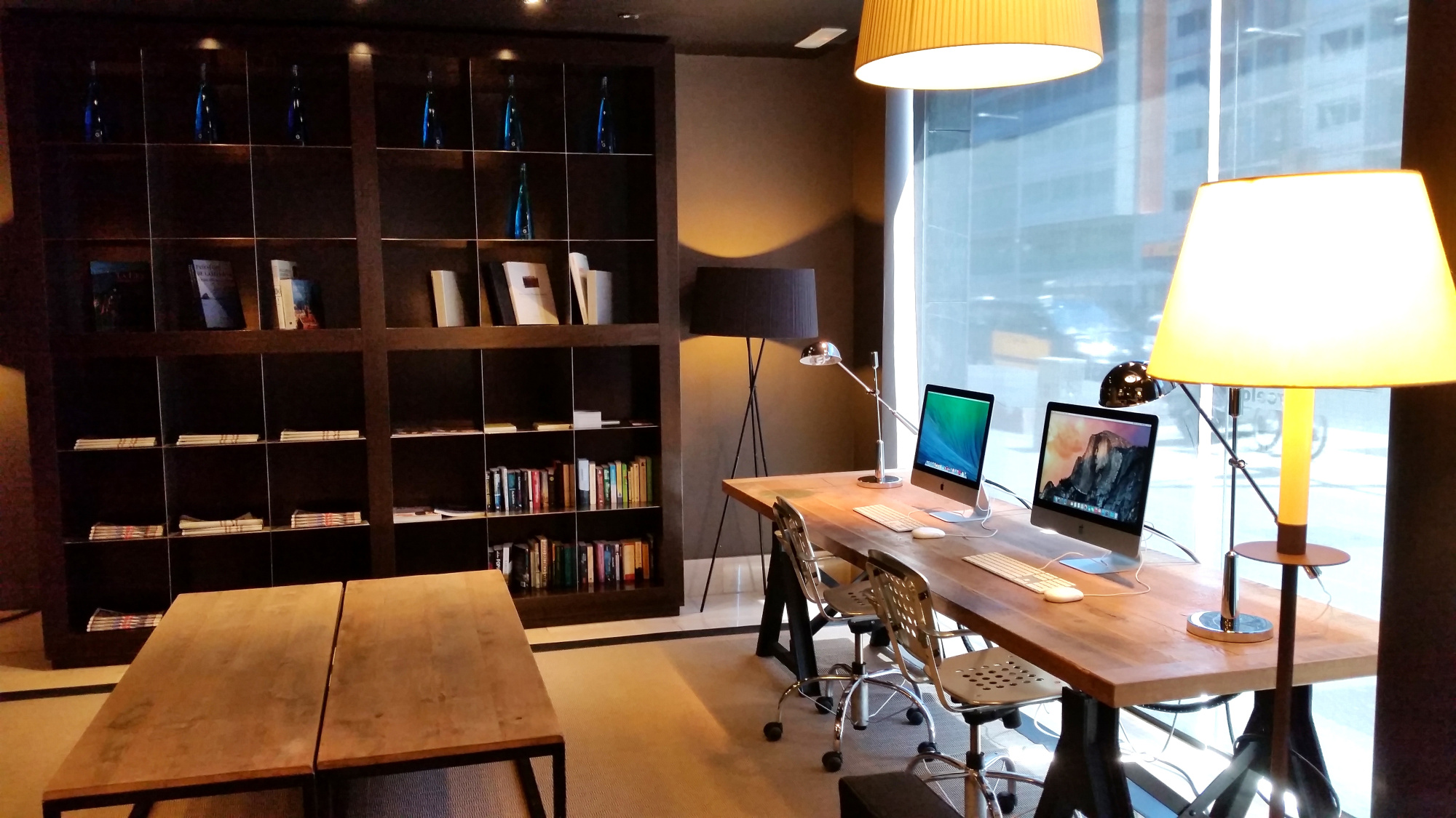 Se ve una habitación interior con escritorio, silla y lámparas, junto a una estantería y una ventana con cortinas.