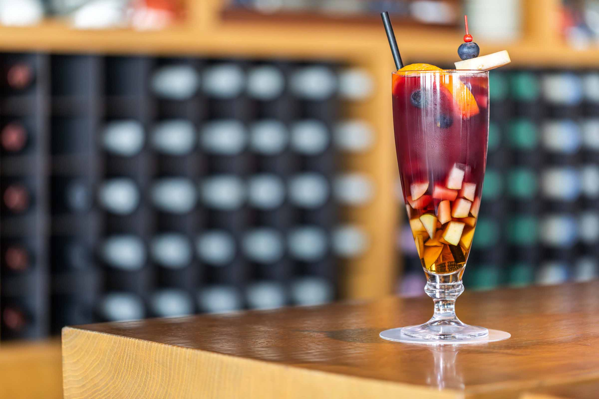 Se ve un cóctel con hielo y una pajita sobre una barra de madera, con botellas desenfocadas al fondo.