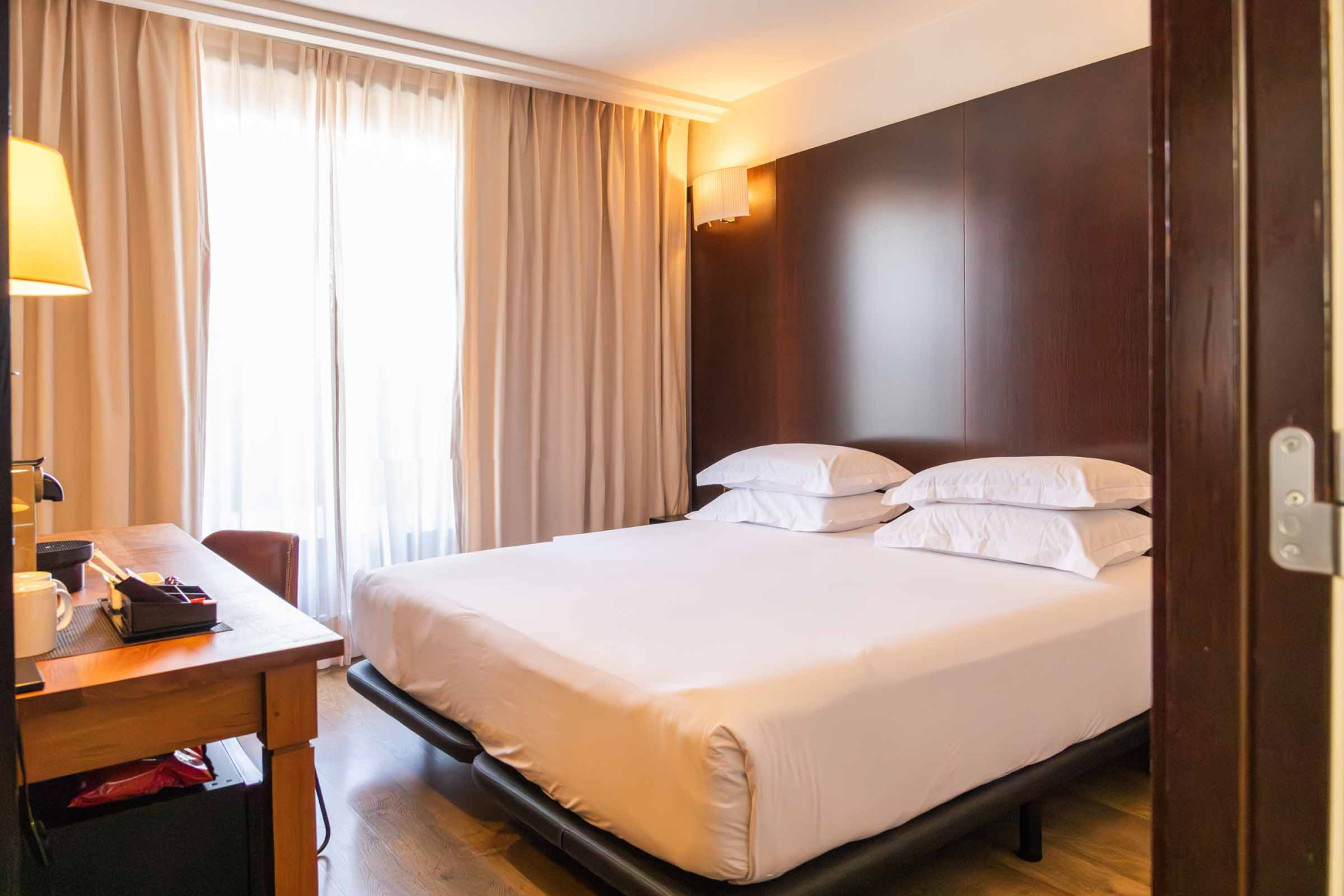 Se ve una cama doble con cojines blancos junto a una mesita de noche y cortinas claras.
