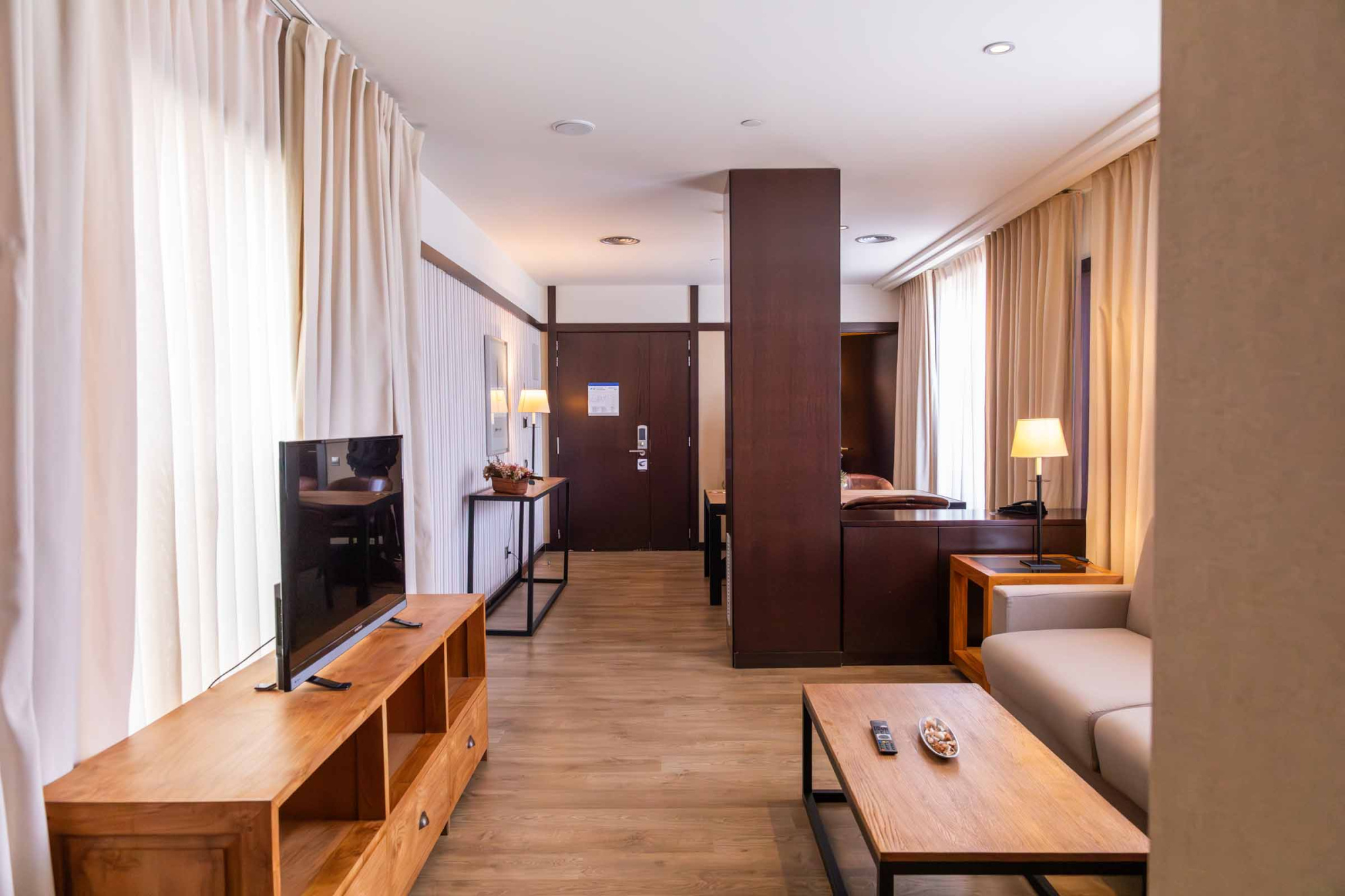 Se ve una sala con sofá beige, mesa baja de madera y televisión junto a cortinas claras.