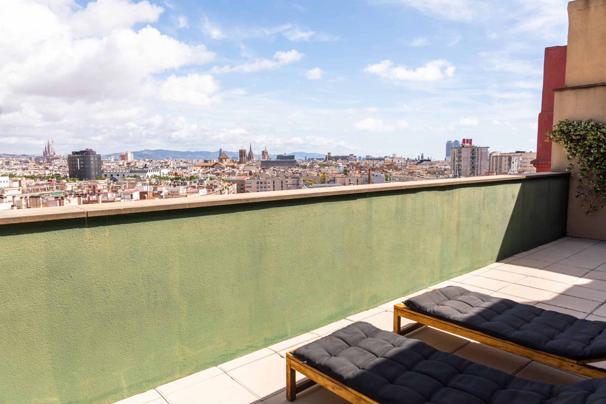Se ve una terraza con tumbonas, una barandilla verde y el skyline urbano al fondo.