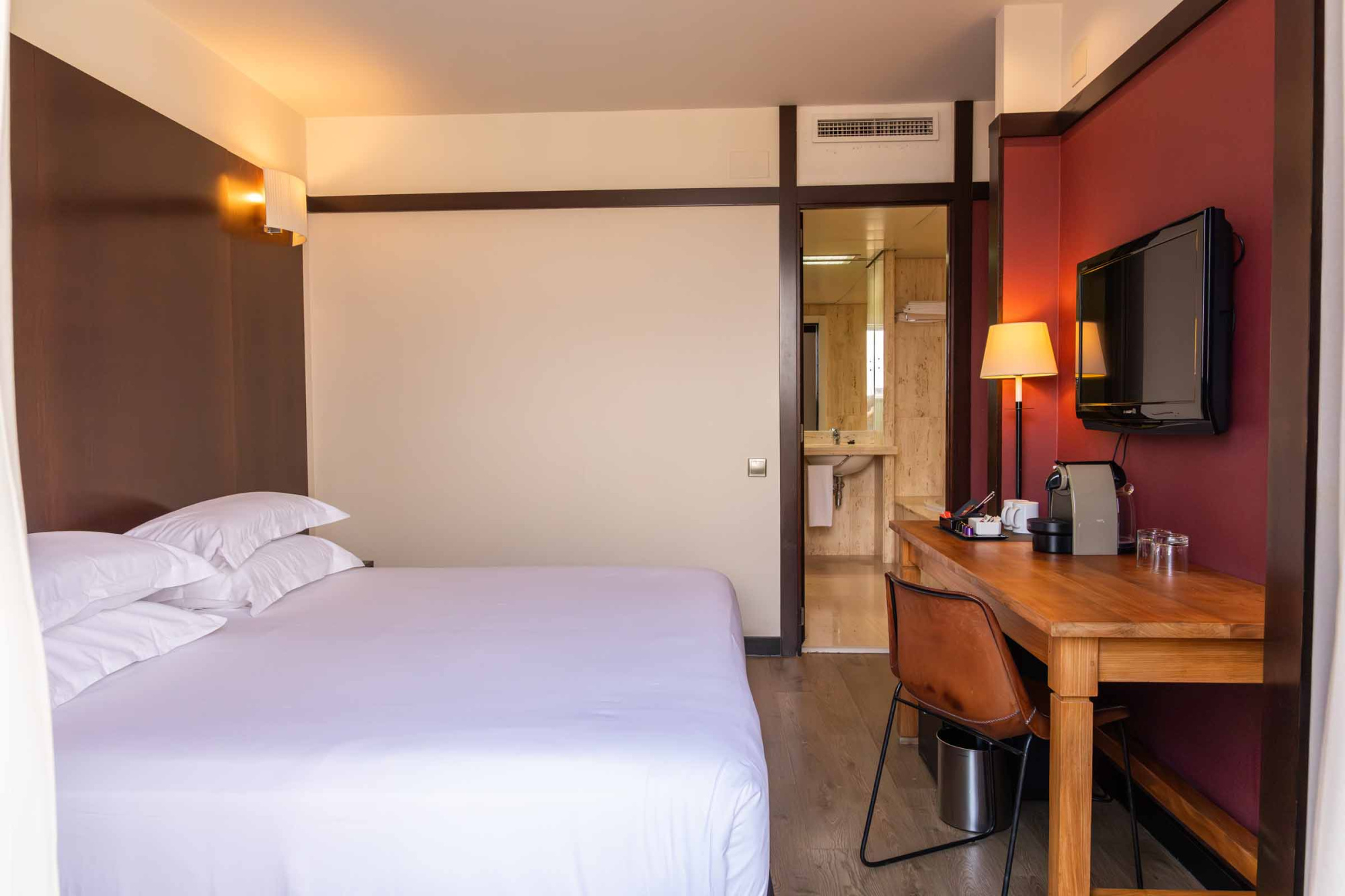 Se ve una cama doble con ropa de cama blanca y un escritorio con silla junto a una pared con iluminación tenue.