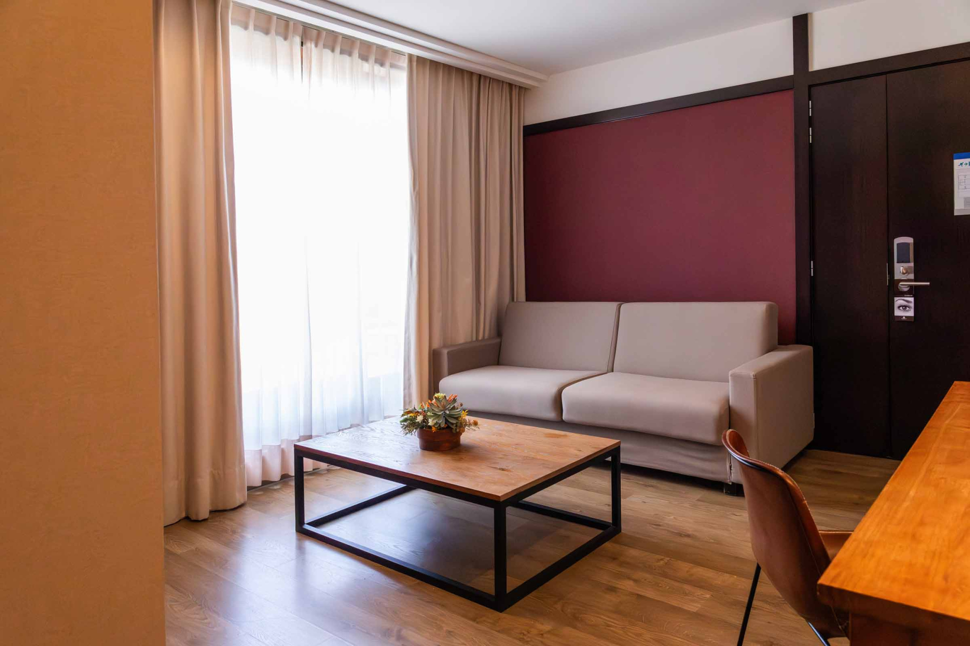 Una habitación luminosa con cama, mesita auxiliar, escritorio y cortinas junto a una ventana.