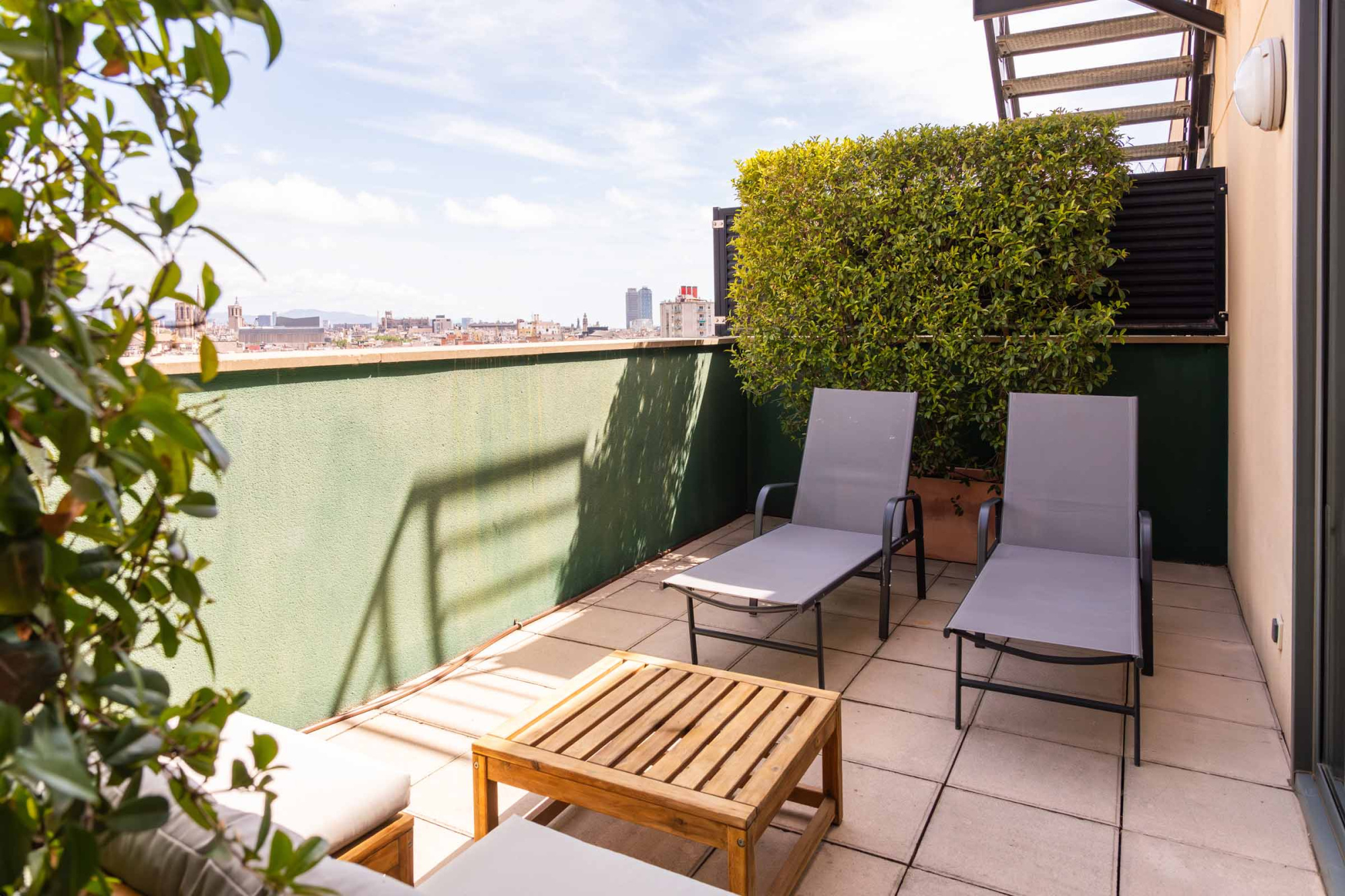 Se ve una terraza exterior con dos tumbonas, una mesa de madera y plantas en jardineras junto a un muro verde.