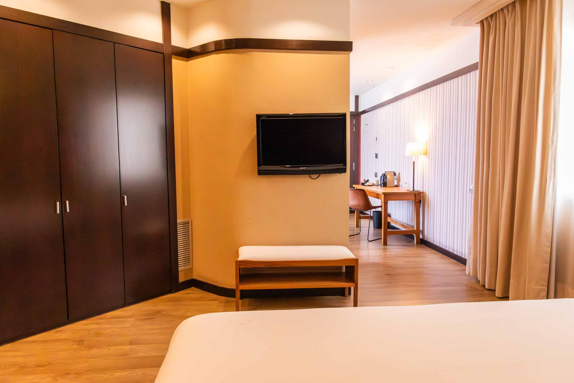 Se ve una cama y un escritorio con televisión en una habitación luminosa con cortinas y suelo de madera.