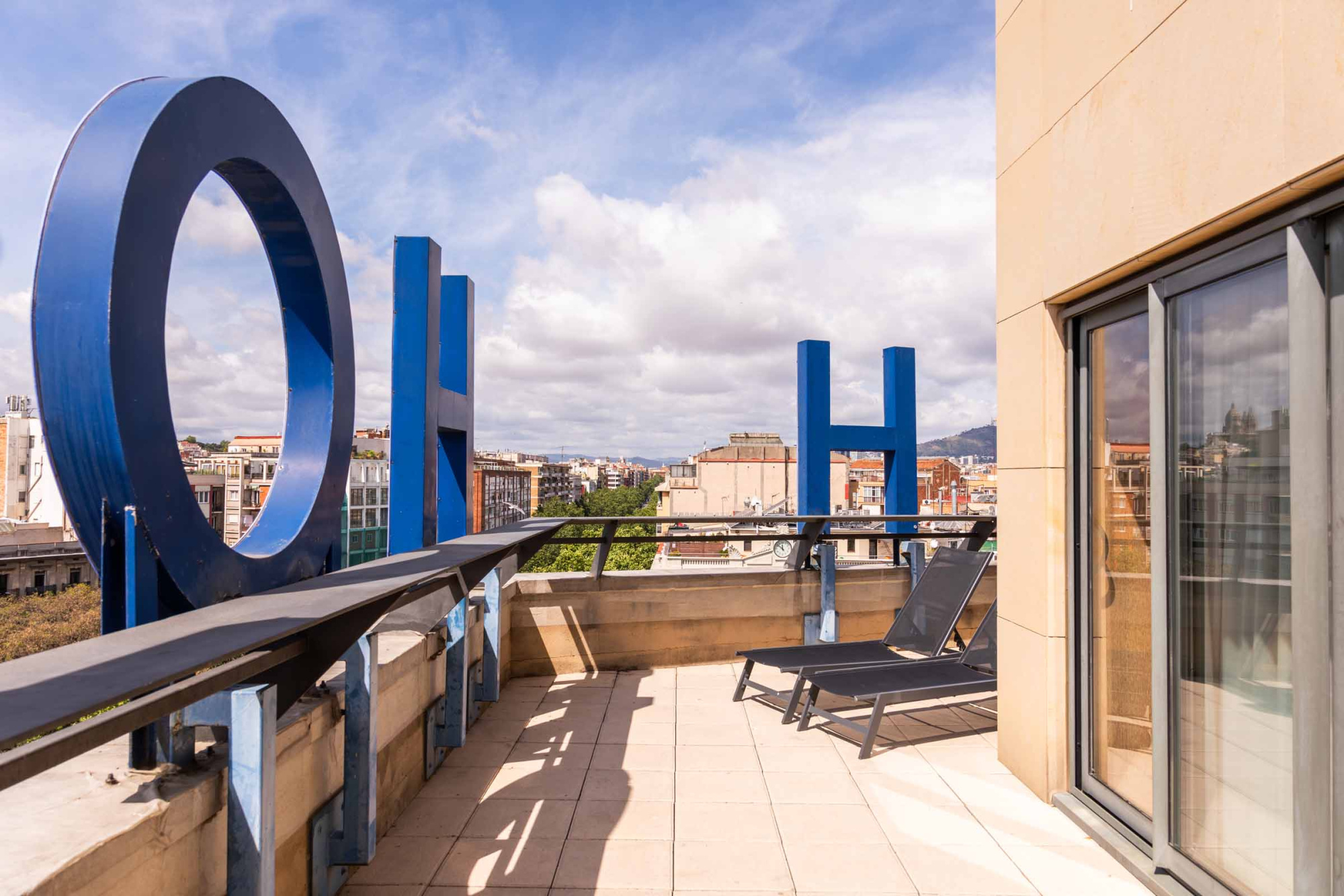 Una terraza exterior con suelo de madera, barandillas y tumbonas, con cielo y edificios al fondo.
