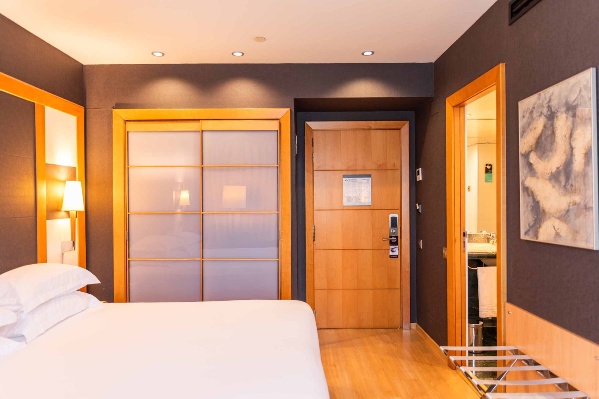 La imagen muestra una cama doble, una puerta y un área de baño con lavabo y espejo.