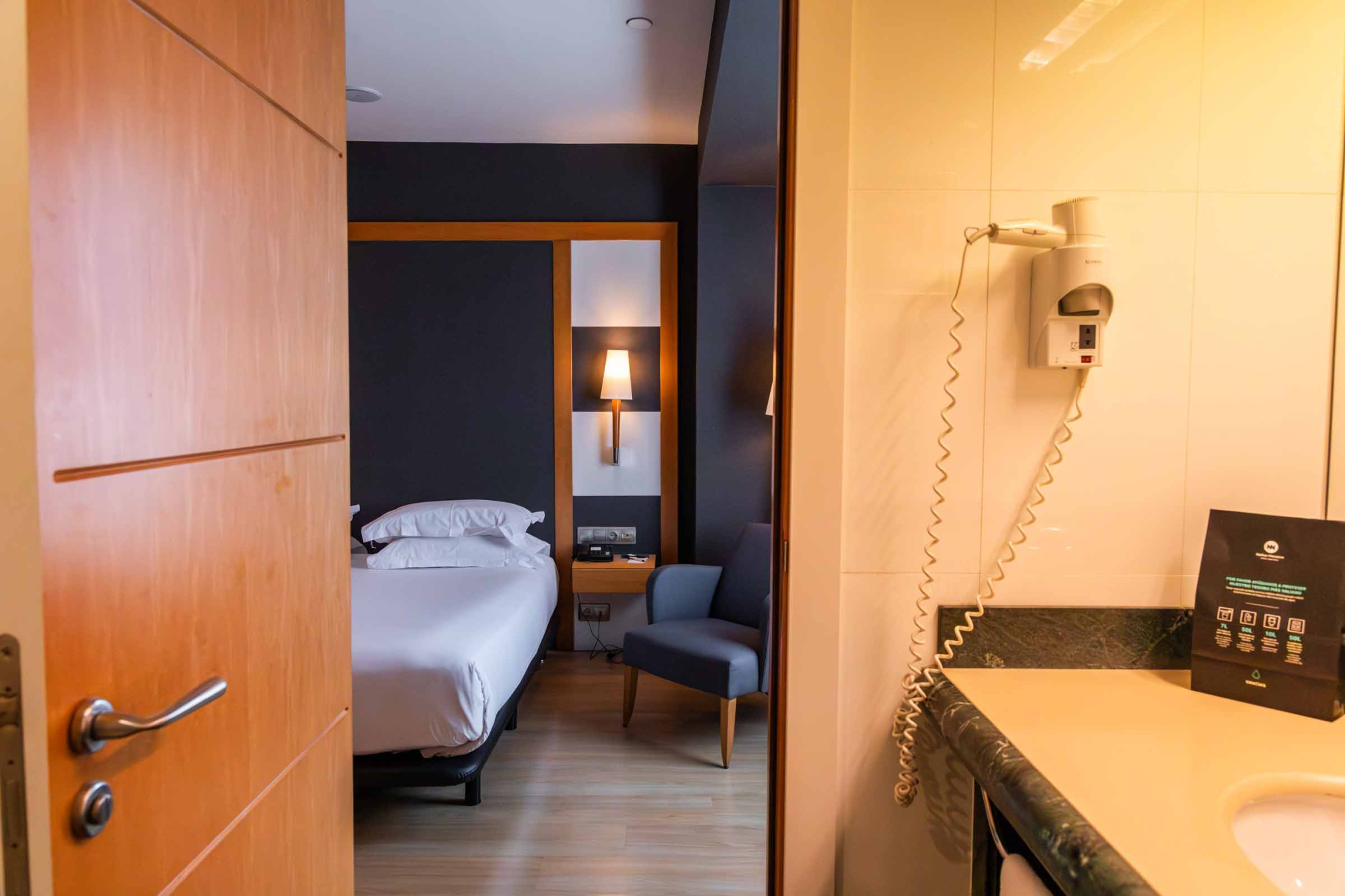 Se ve una puerta abierta hacia una habitación con una cama y un baño con lavabo y espejo al fondo.