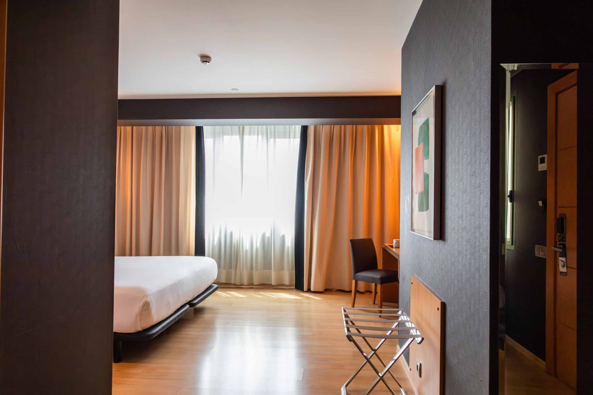 Se ve un dormitorio con una cama, cortinas y una mesa con silla junto a una pared oscura.
