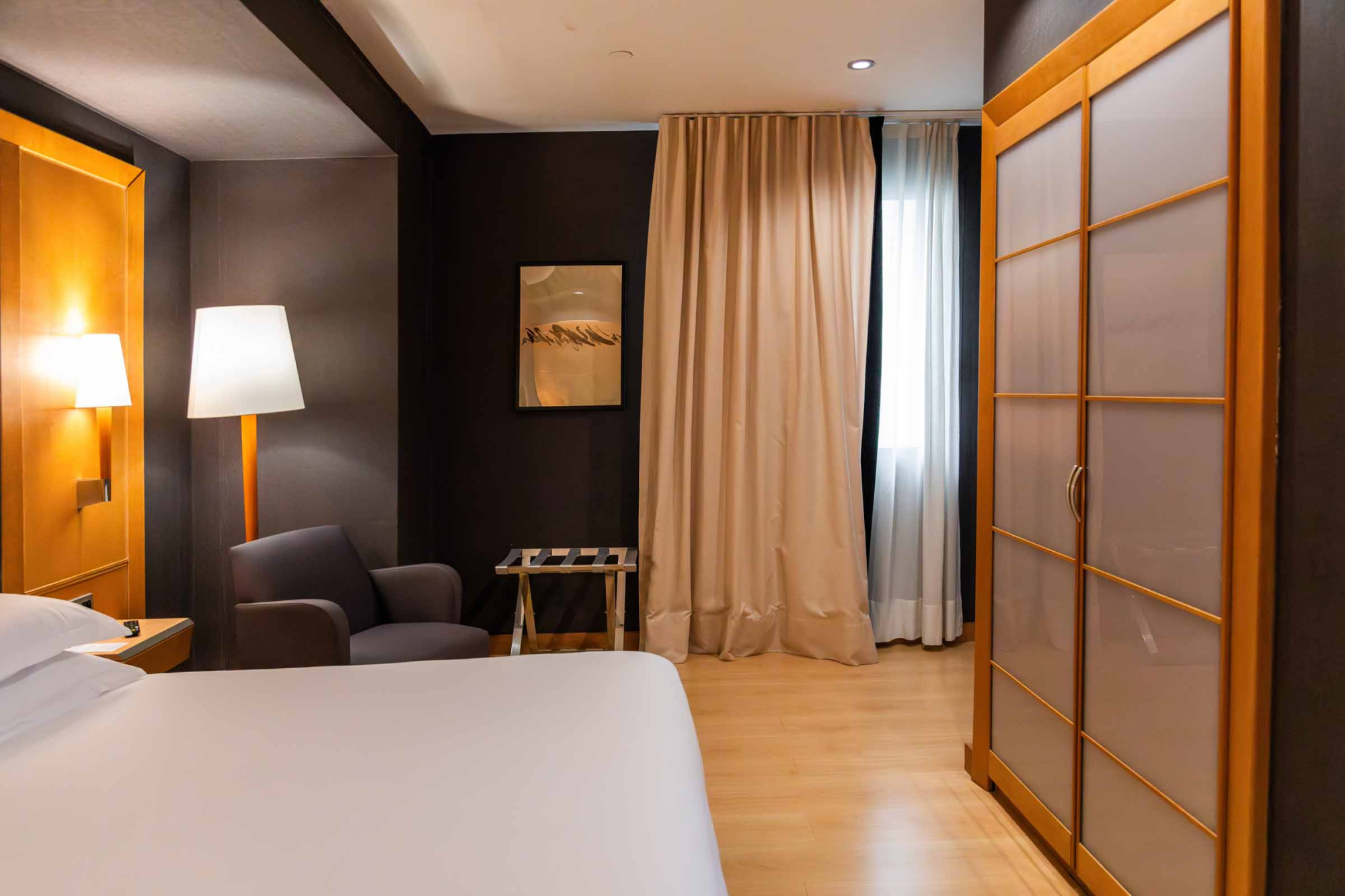 Se ve una cama con ropa de cama blanca, una silla junto a una lámpara y cortinas delante de una ventana.