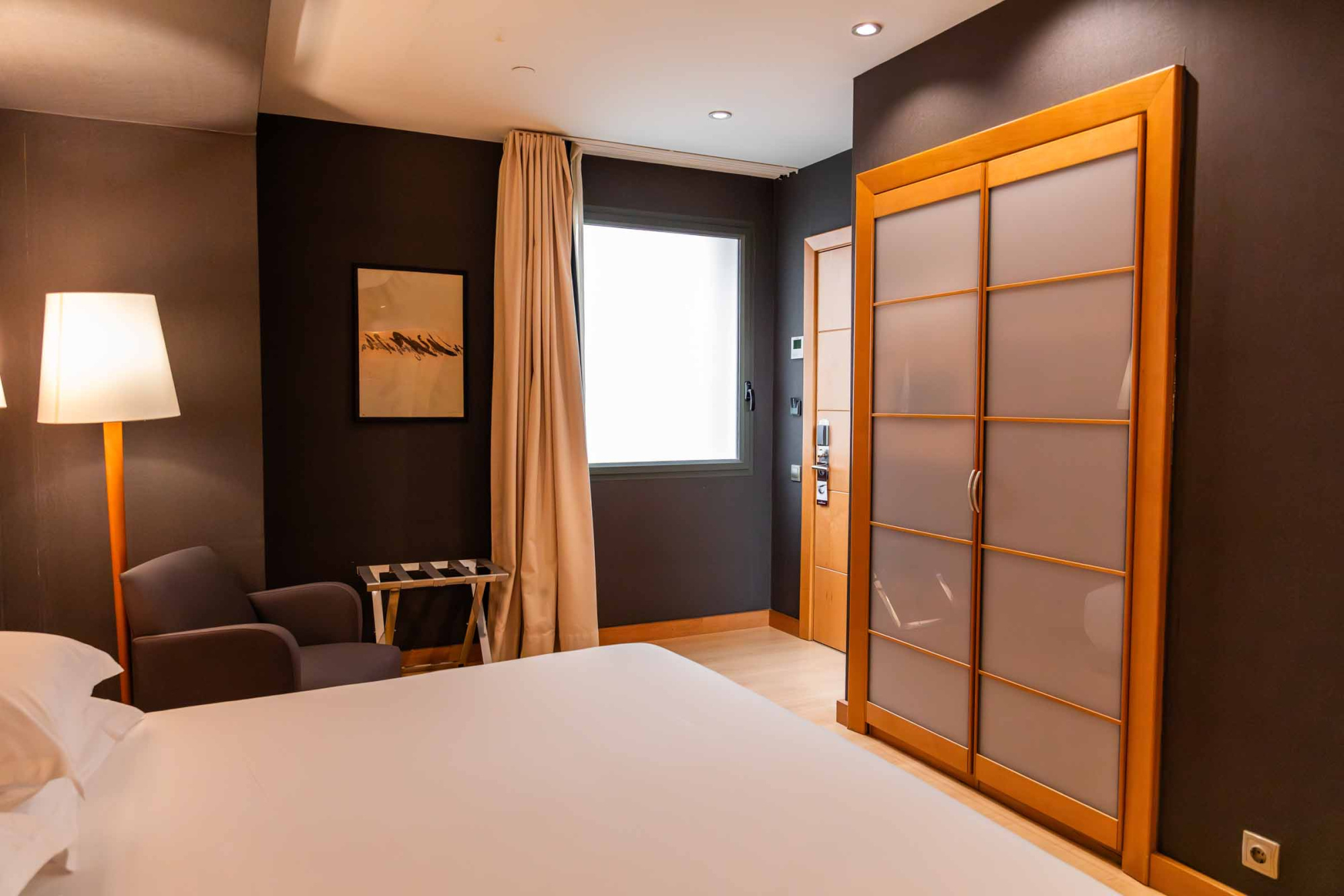 Se ve una cama, una lámpara, un cuadro y una puerta corredera con paneles en una habitación con ventana.