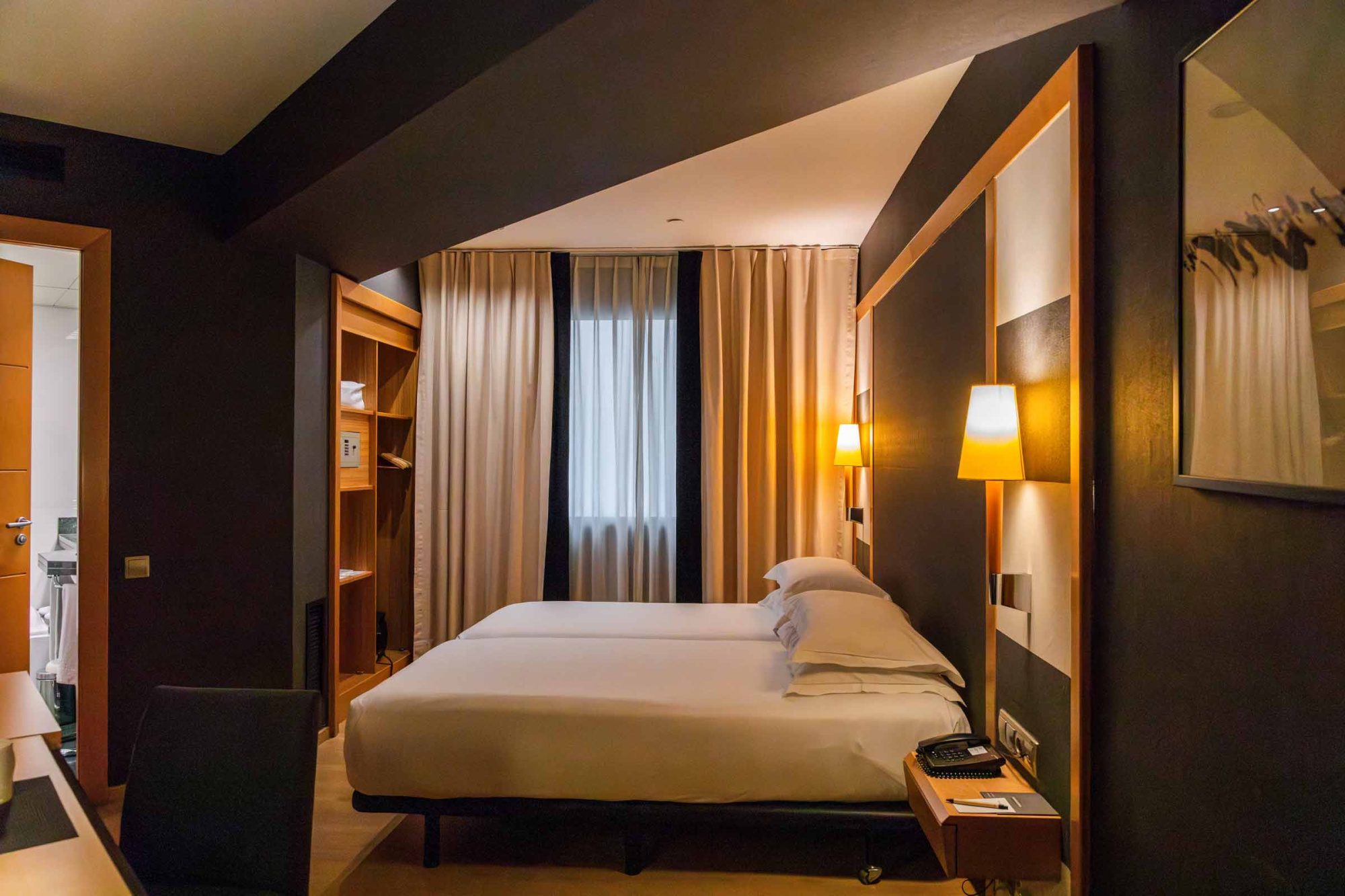 Una cama doble con ropa de cama blanca se ve junto a lámparas de pie y cortinas traslúcidas.