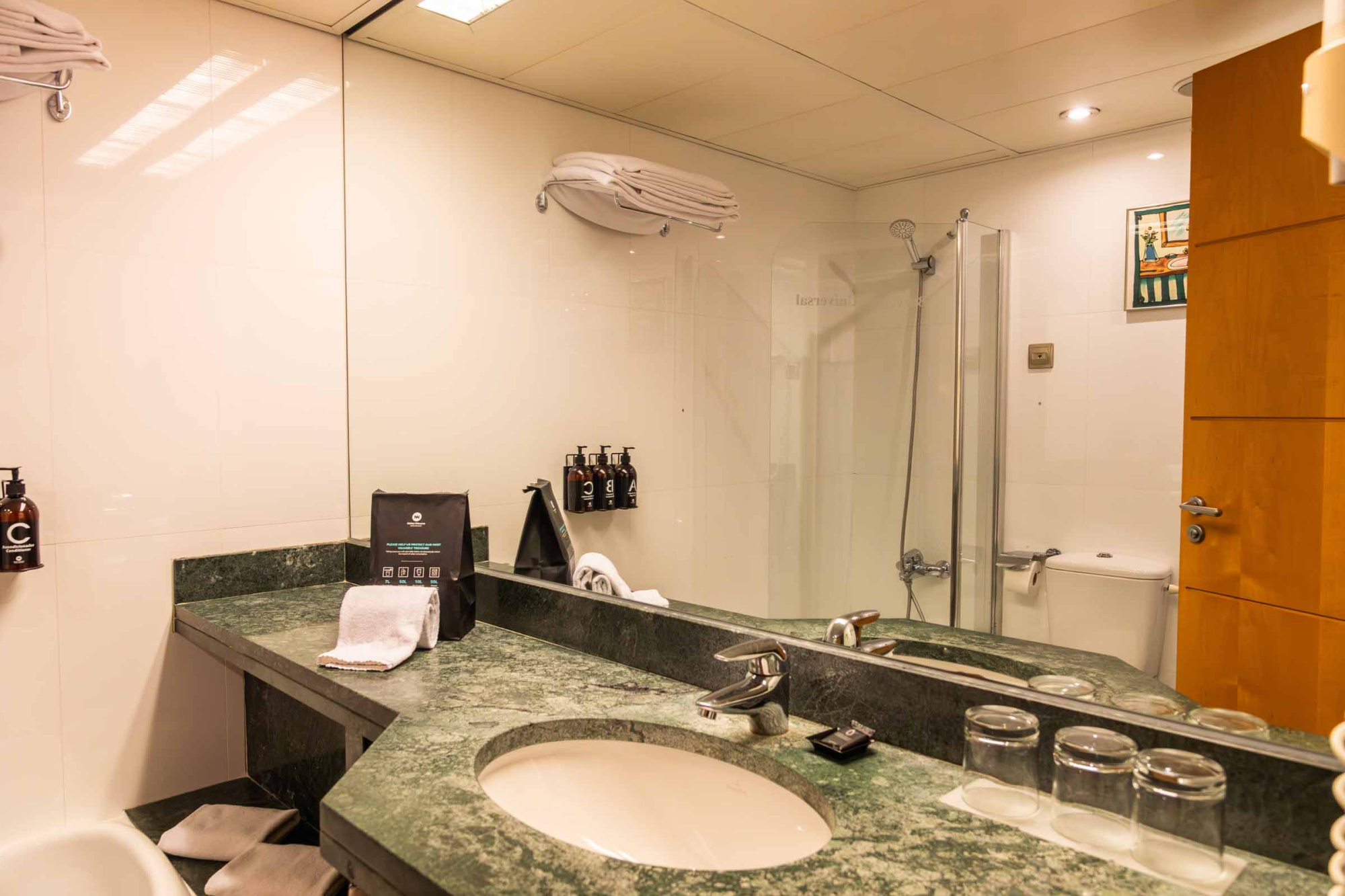 El baño muestra un lavabo con grifería, espejo, ducha y dispensadores sobre la encimera.