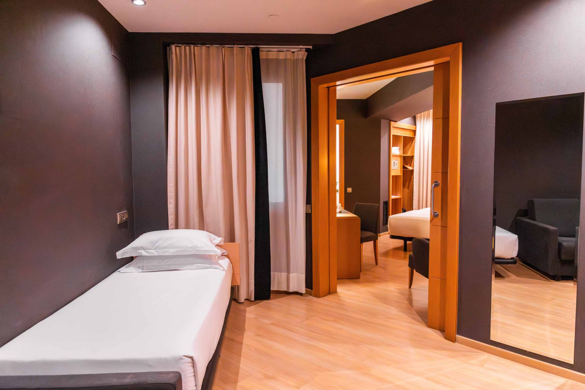 La imagen muestra una cama doble, cortinas y una zona de estar con sillón y mesa a través de una abertura interior.