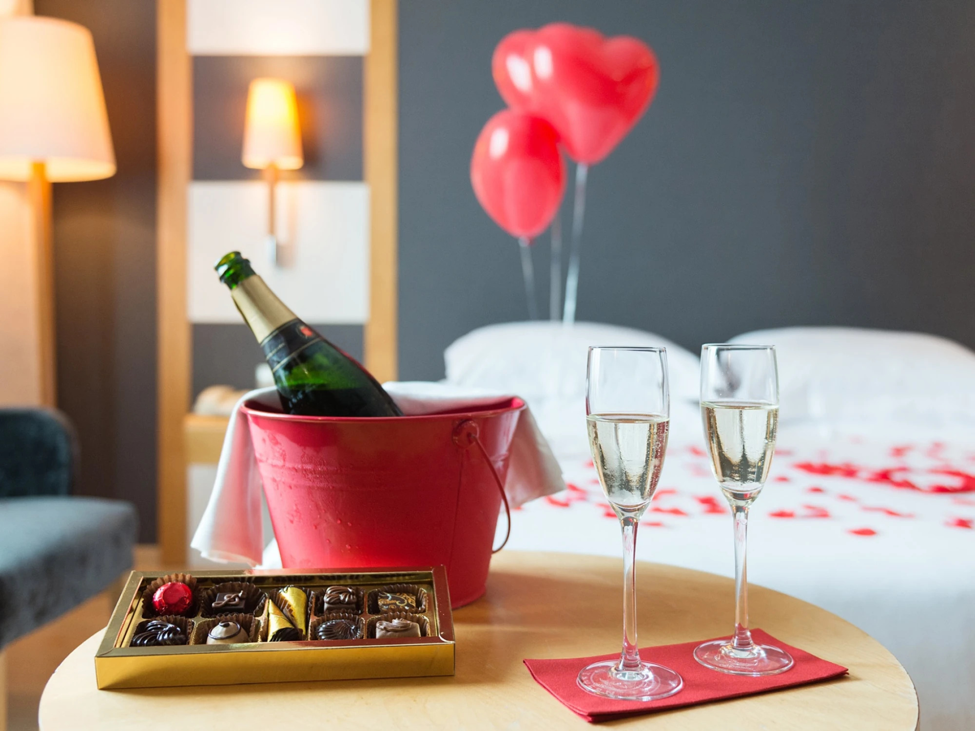 Una mesa con botella, copas y decoración romántica sobre una cama, con una lámpara encendida al fondo.