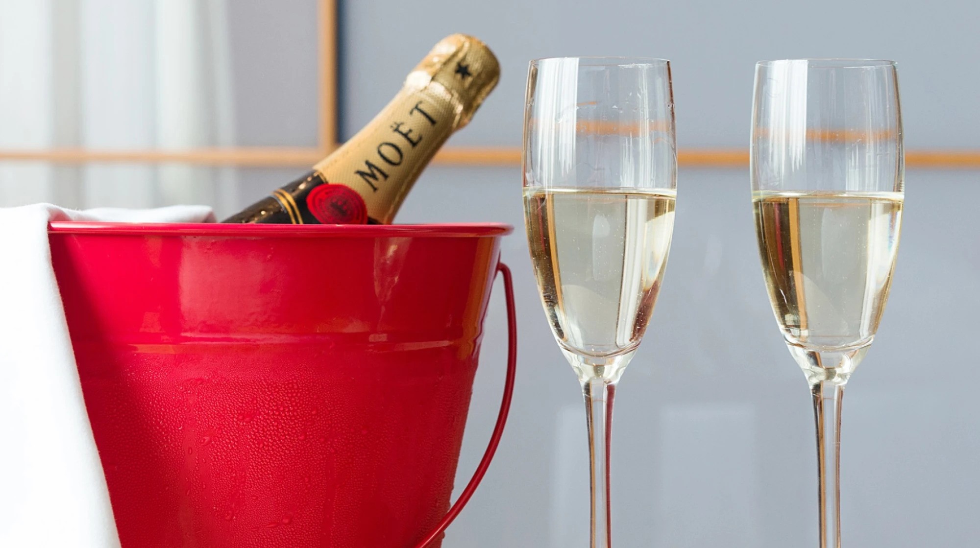 Una botella con etiqueta y copas de champán se muestran sobre una mesa con un cubo rojo al frente.