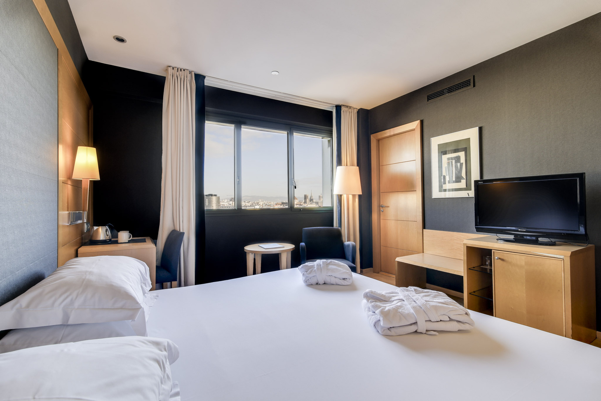 La imagen muestra una habitación con dos camas, una televisión, una mesa auxiliar y luz natural entrando por una ventana.