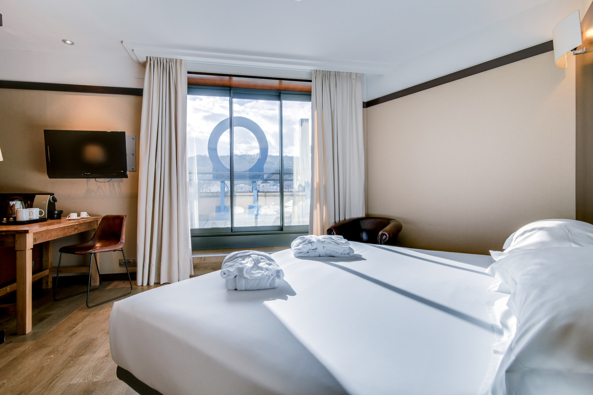 En la imagen se ve una cama doble con almohadas y una ventana con cortinas junto a una pequeña mesa y una lámpara.