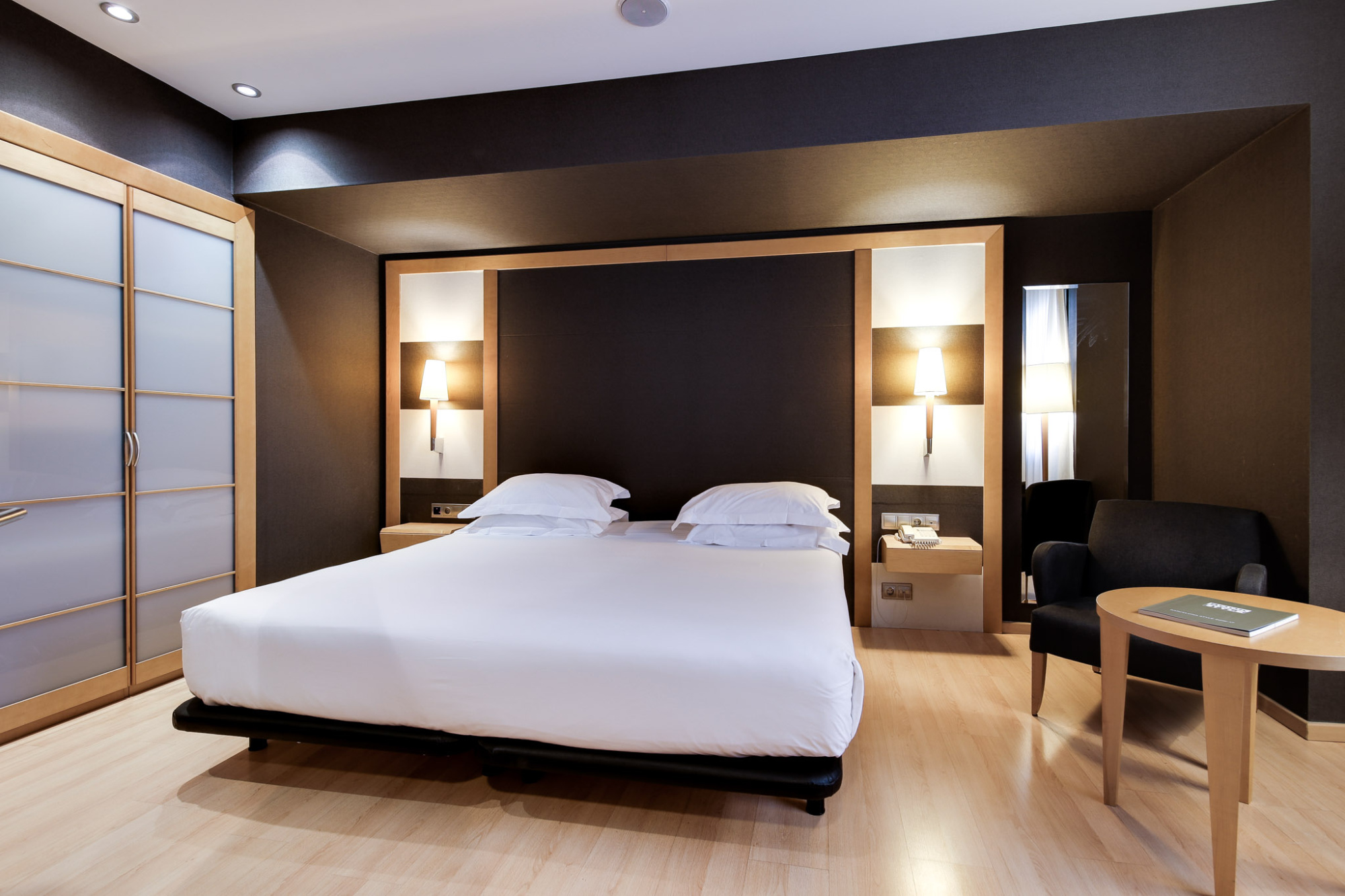 La imagen muestra una cama doble con ropa de cama blanca y un armario a la izquierda, junto a una zona de estar con mesa y butaca.