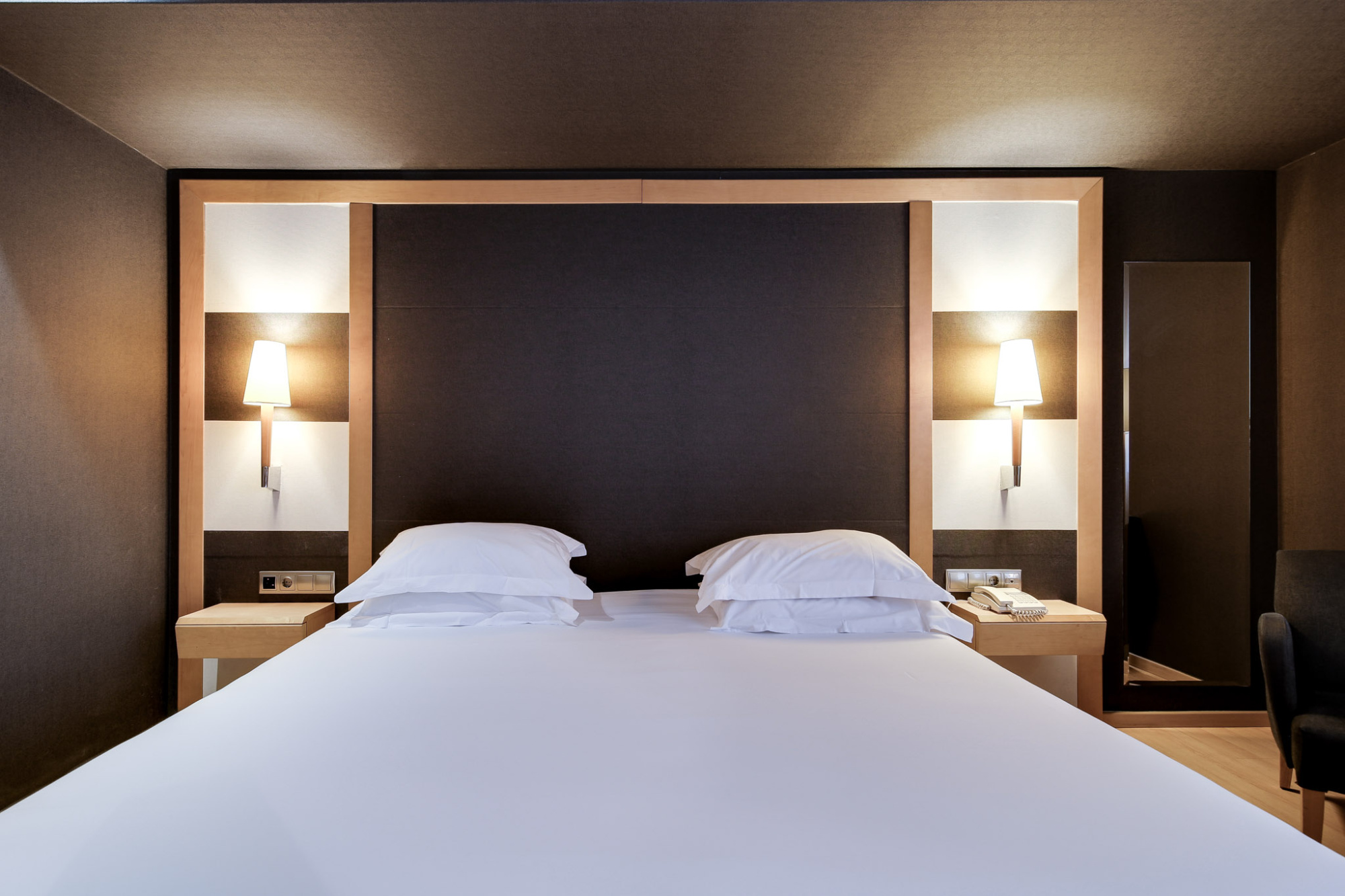 Una habitación moderna muestra una cama doble con dos mesitas y lámparas de pared encendidas.