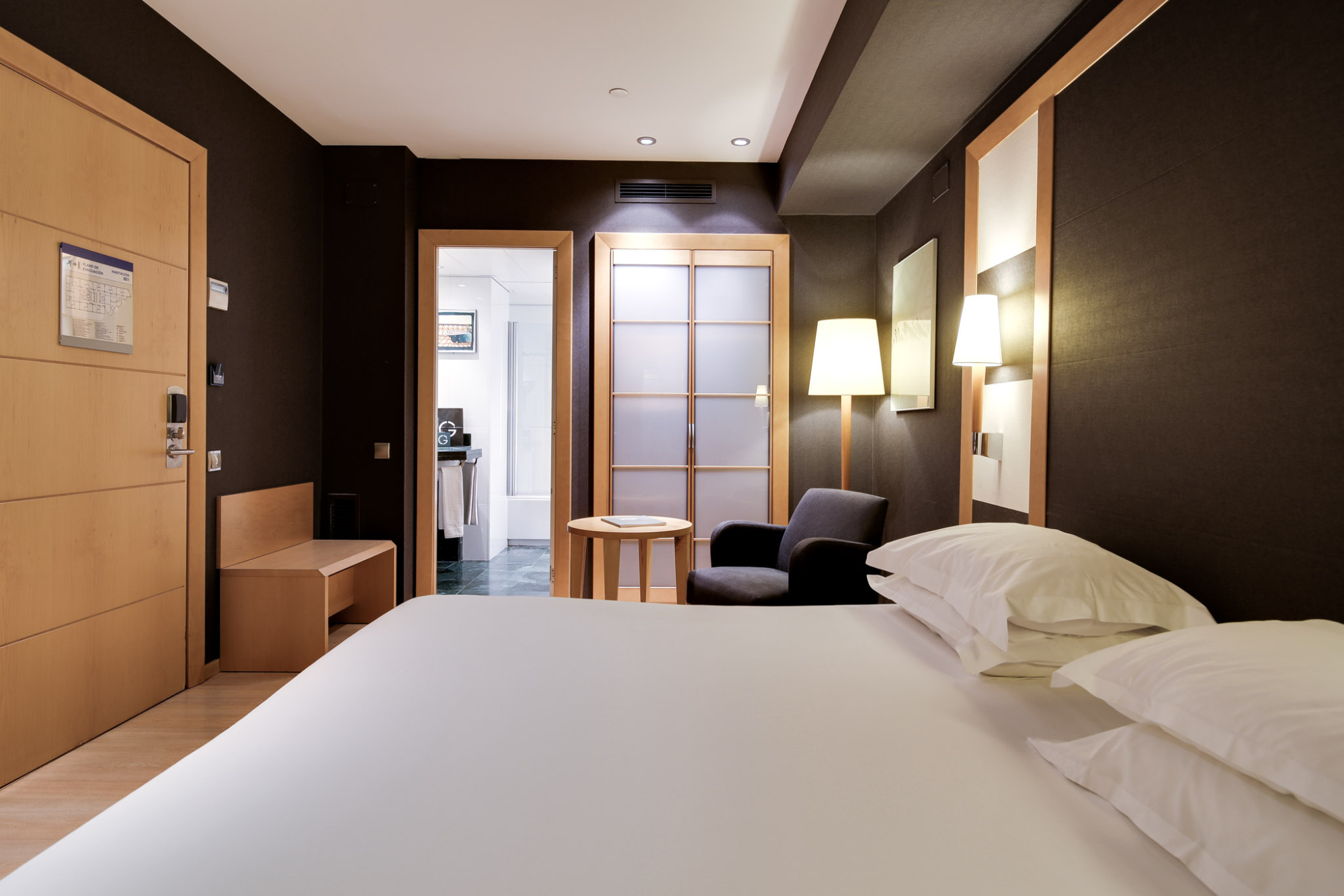 Se ve una cama con ropa de cama blanca y una silla junto a una mesa, con luz de pared y puerta al fondo.